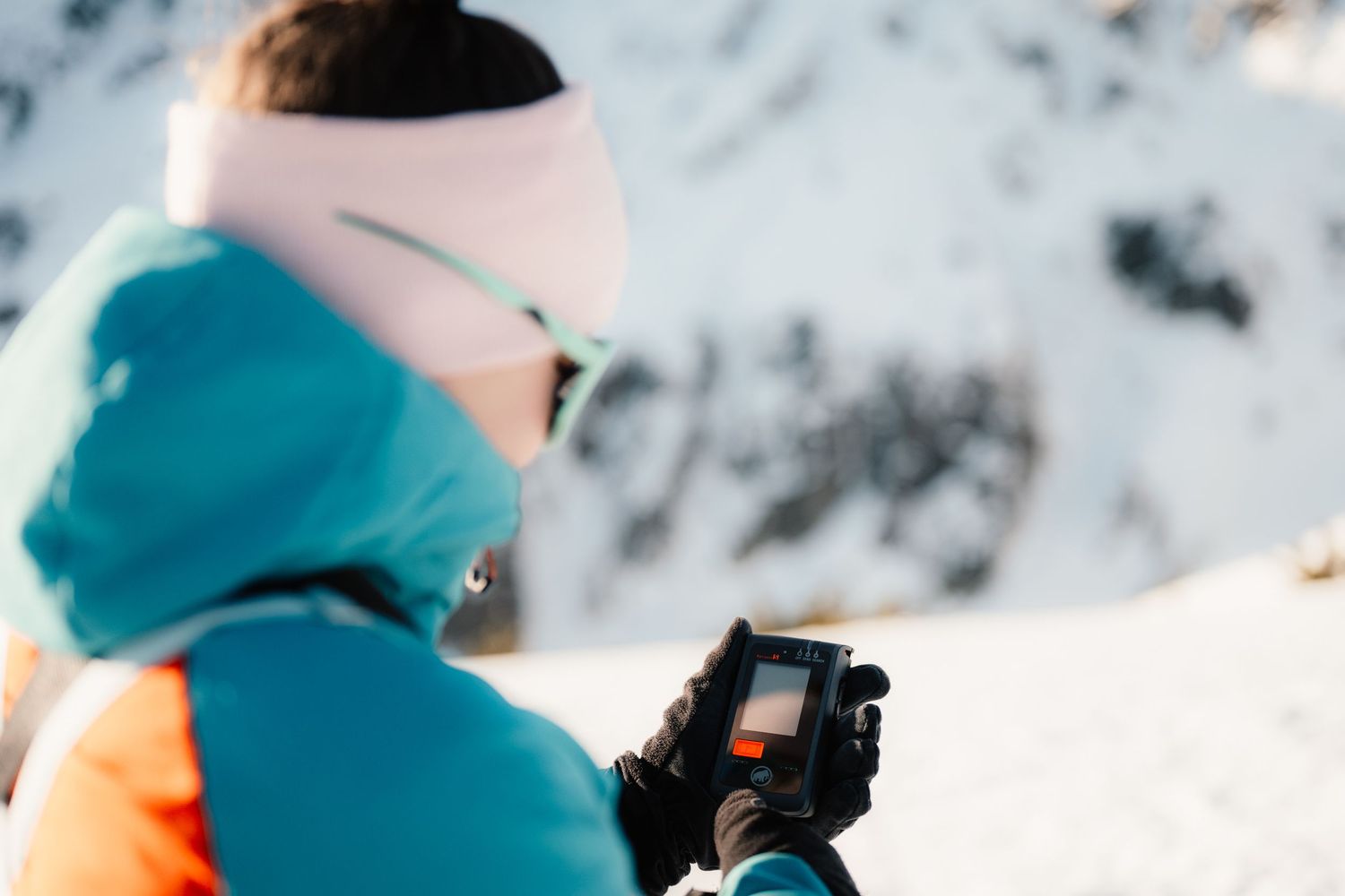 Female backcountry skier in pink beanie checking avalanche rescue device on bright snowy mountain slope.