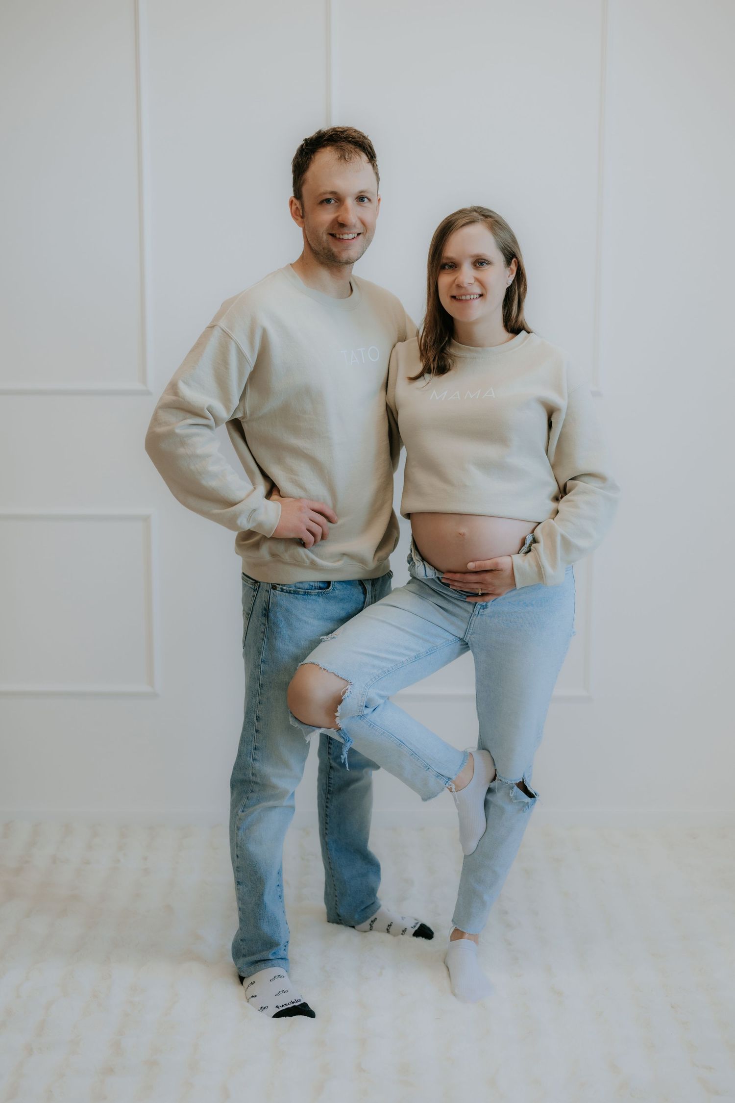 Smiling pregnant couple in matching beige sweatshirts and ripped jeans pose against white wall.