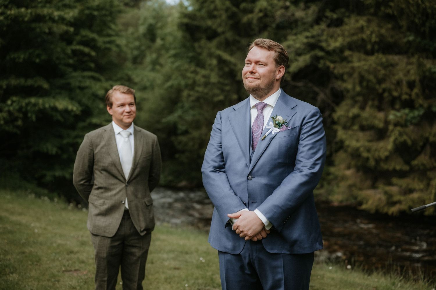 Two groomsmen in suits stand outdoors near evergreen trees at a wedding ceremony.