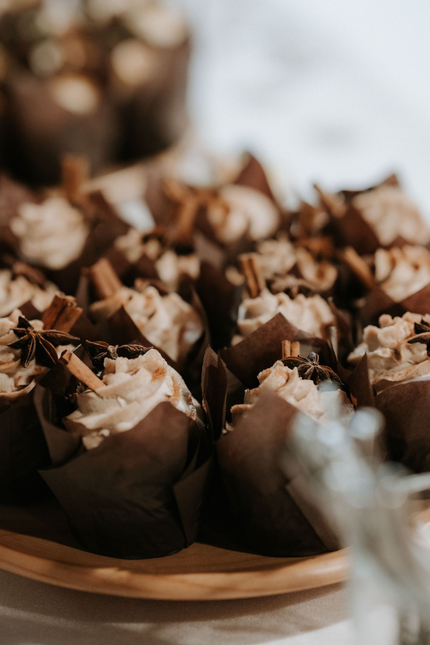 Elegant cupcakes in brown paper wrappers topped with cream frosting and cinnamon sticks on a wooden plate.