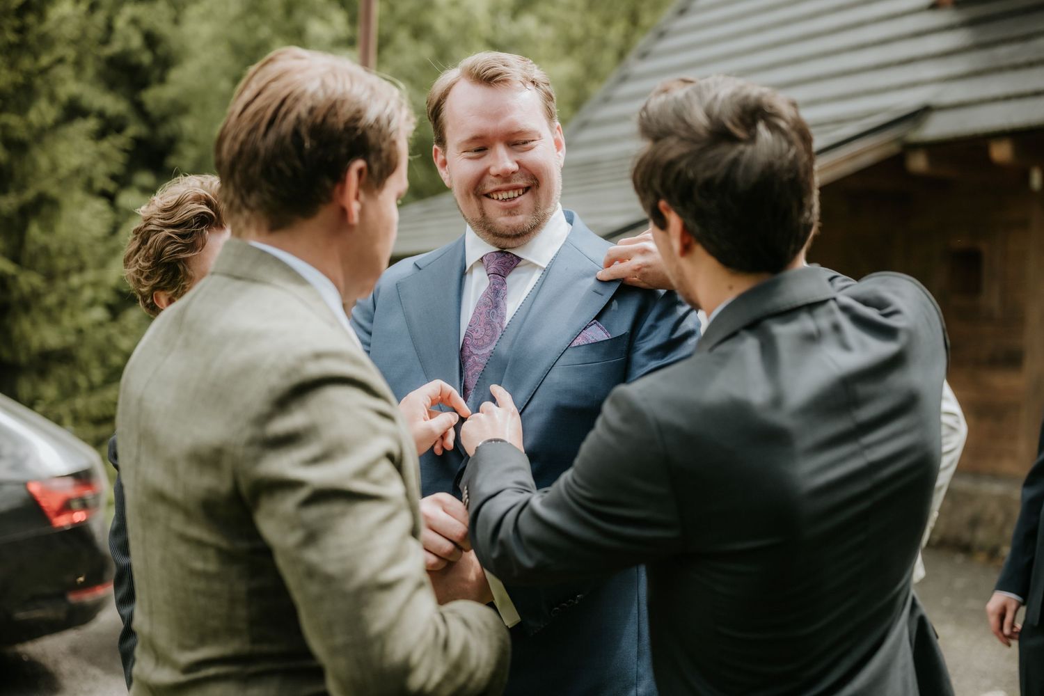 Groom in blue suit laughing with groomsmen while getting boutonniere pinned at outdoor wedding.