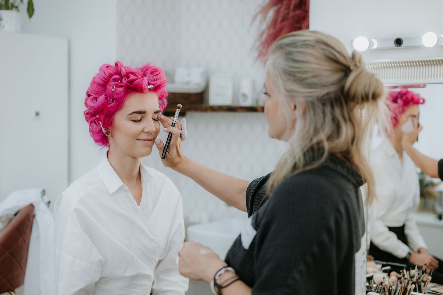 A makeup artist applies foundation to a woman with pink hair rollers in a bright salon with mirror lighting.