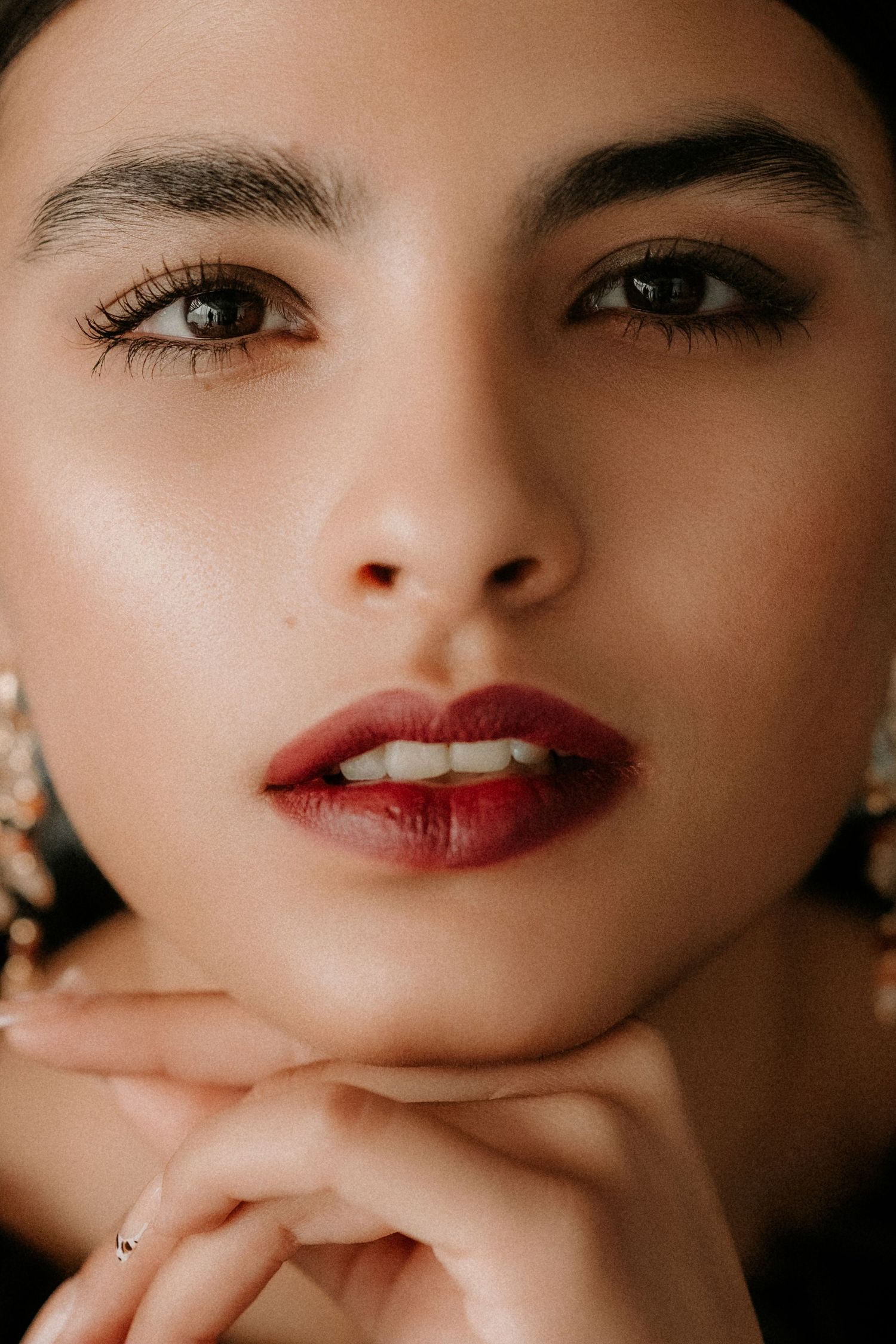 Close-up portrait of a young woman with bold brows, deep red lips, and chandelier earrings resting chin on finger.