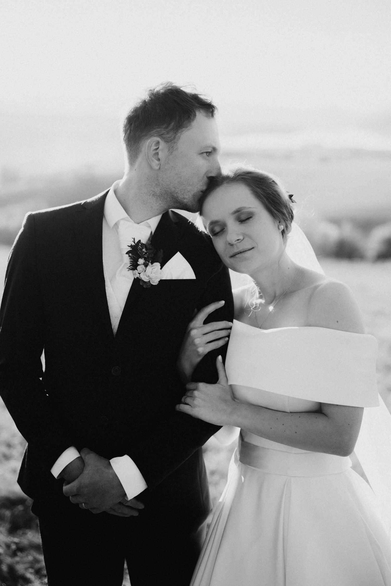 Black and white wedding photo of groom kissing bride on forehead as she leans against him outdoors.
