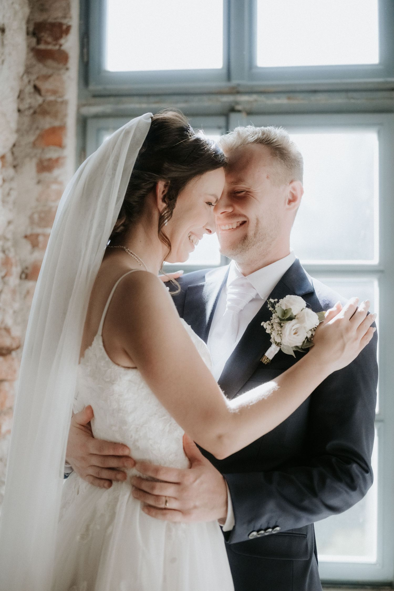 A smiling bride and groom embrace tenderly indoors, with the bride wearing a veil and the groom a boutonniere.