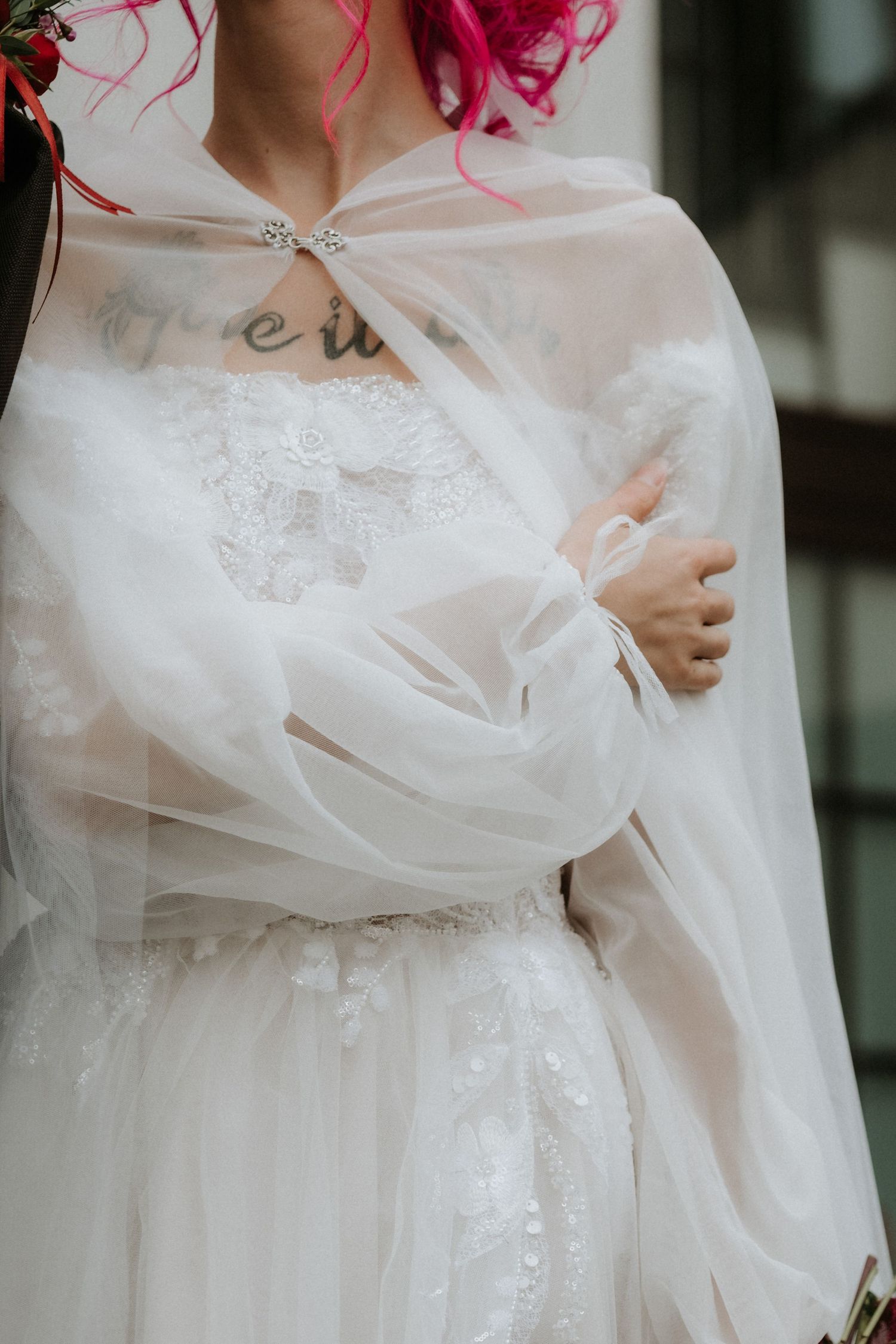 Pink-haired bride in white lace wedding dress wearing sheer tulle capelet outdoors.