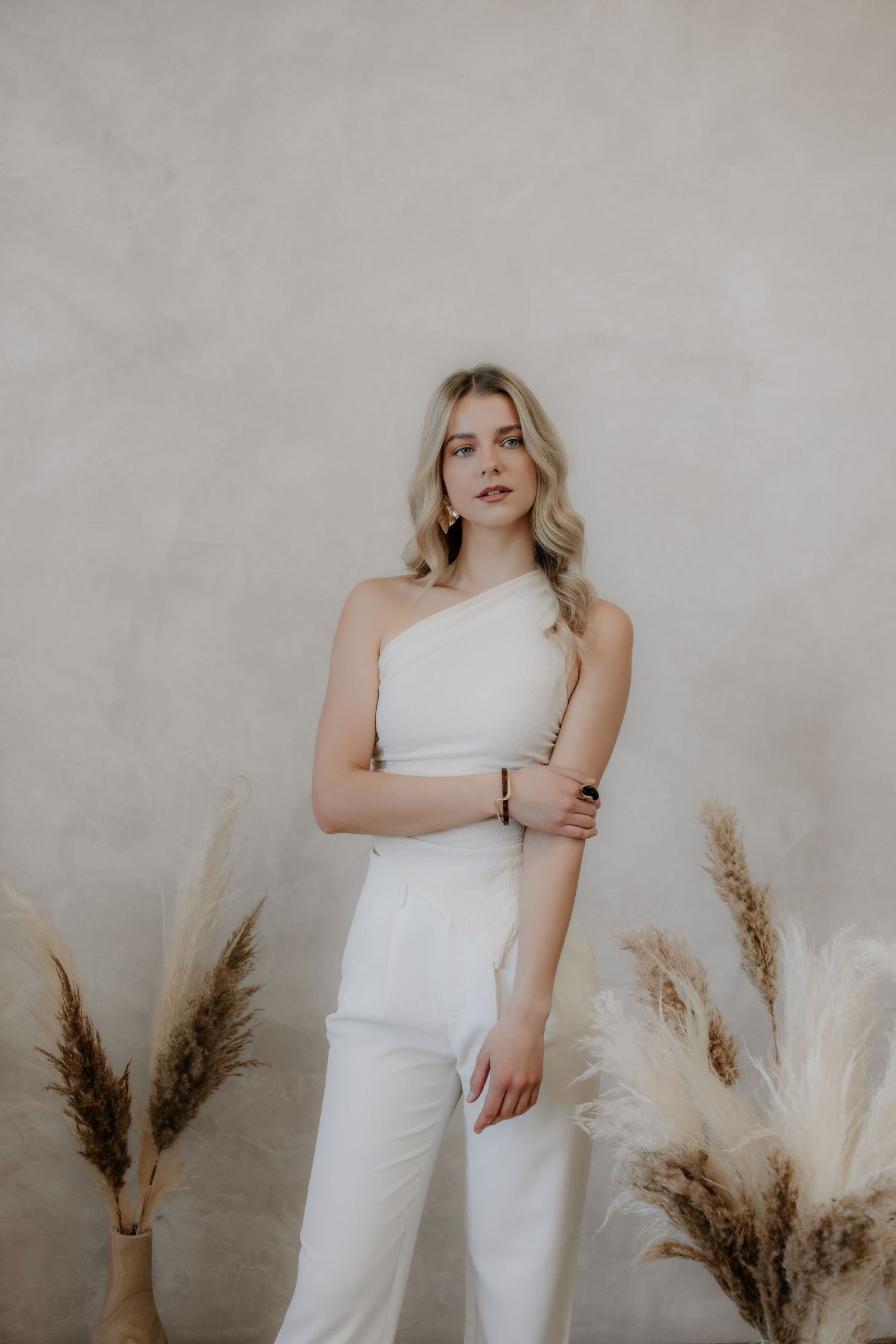 Blonde woman in white one-shoulder jumpsuit posing with dried pampas grass against neutral wall.