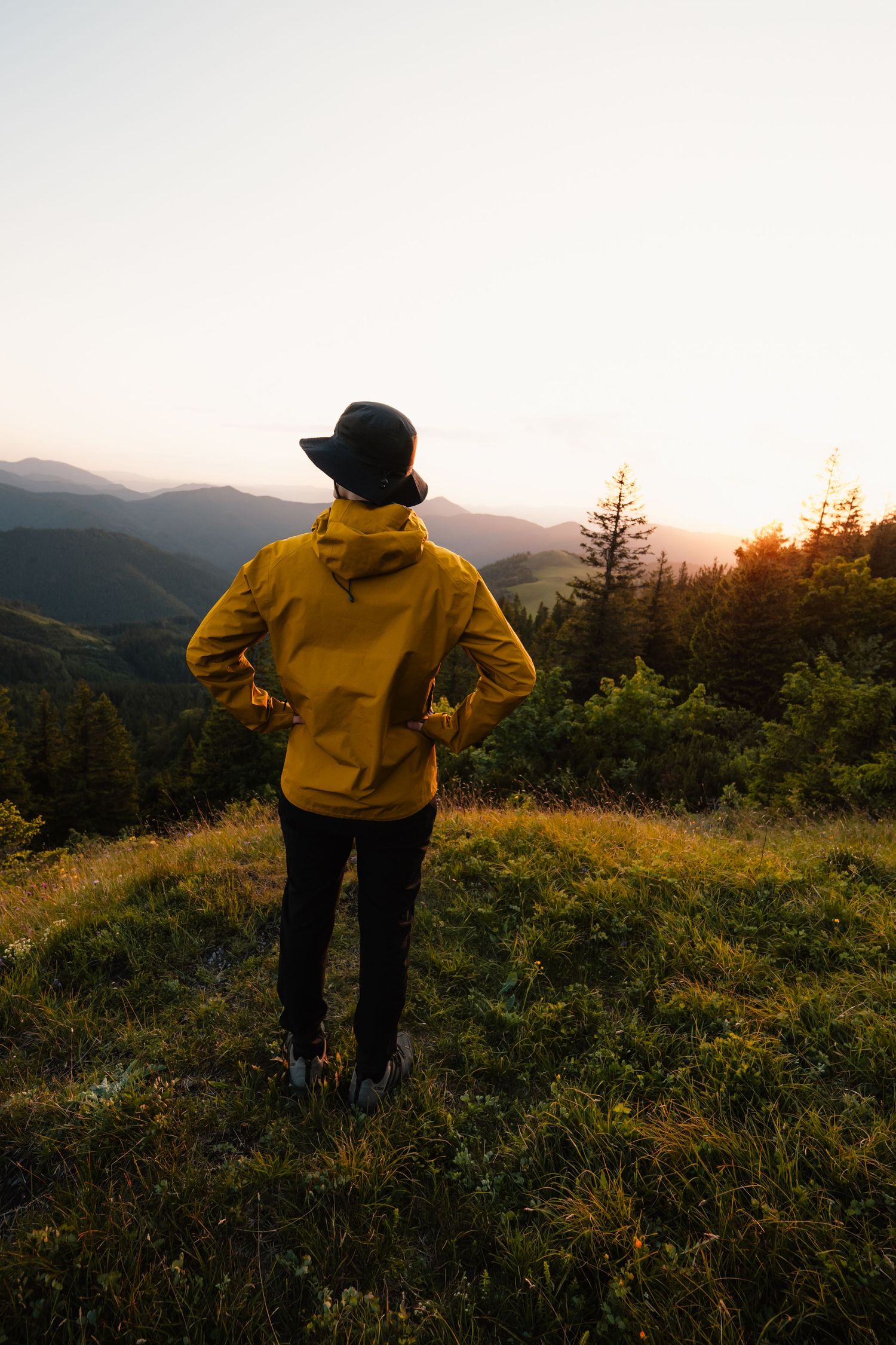 A person in a yellow rain jacket and black hat stands on a mountain hilltop admiring the sunset view.