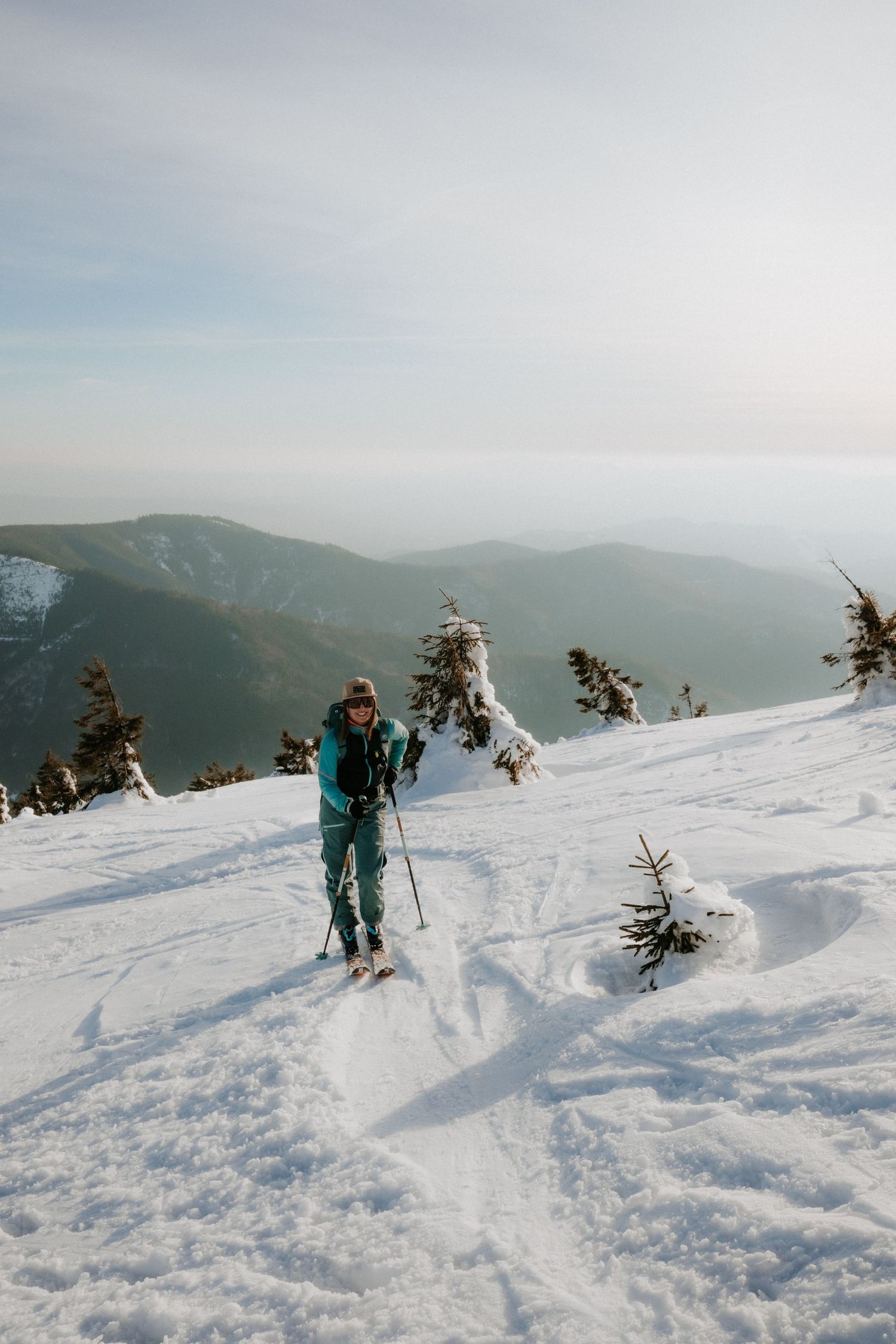 Female backcountry skier in teal suit ascending snowy peak with panoramic mountain valley views.