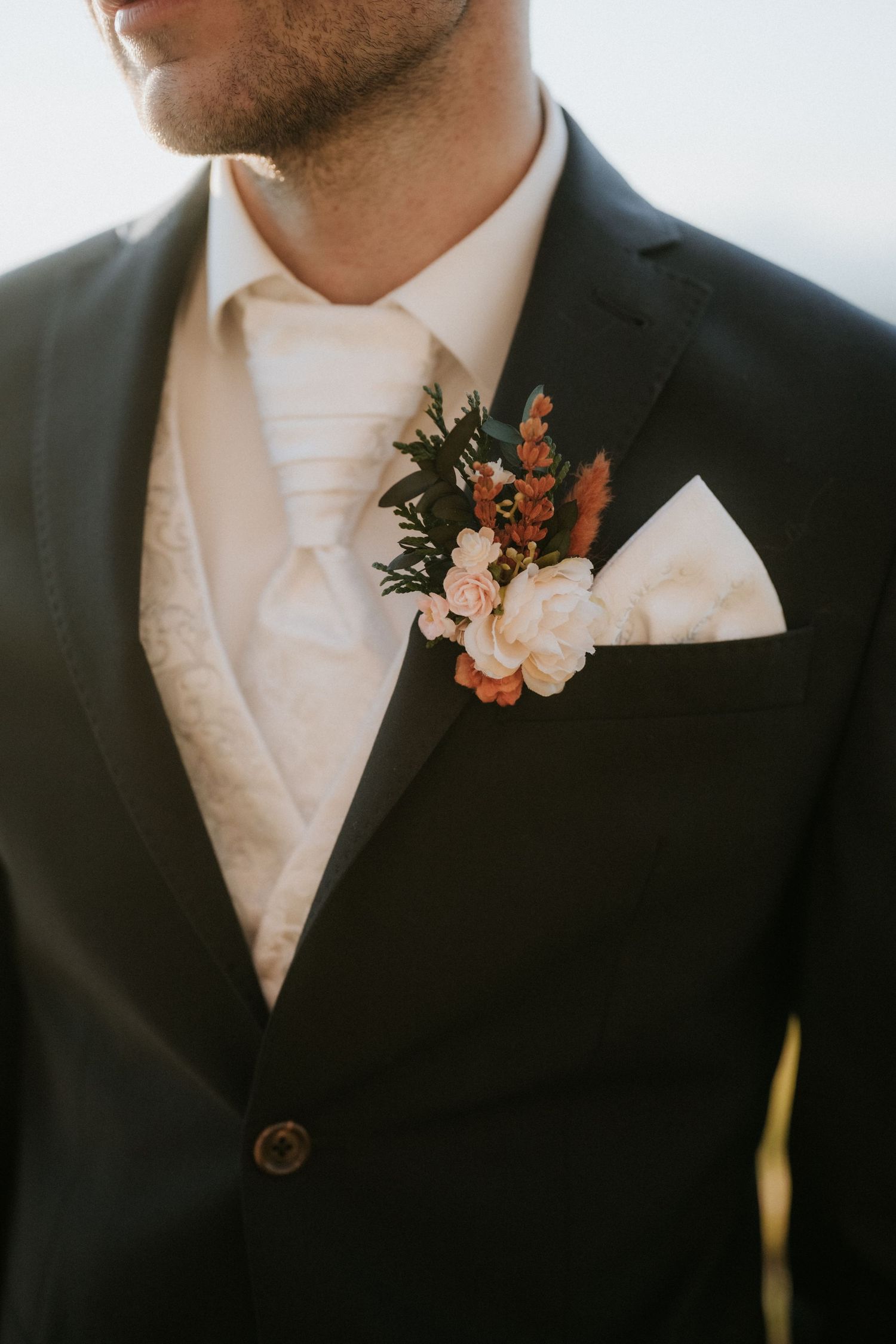 Groom in dark suit with floral boutonniere featuring white and terracotta blooms, ivory tie and pocket square.