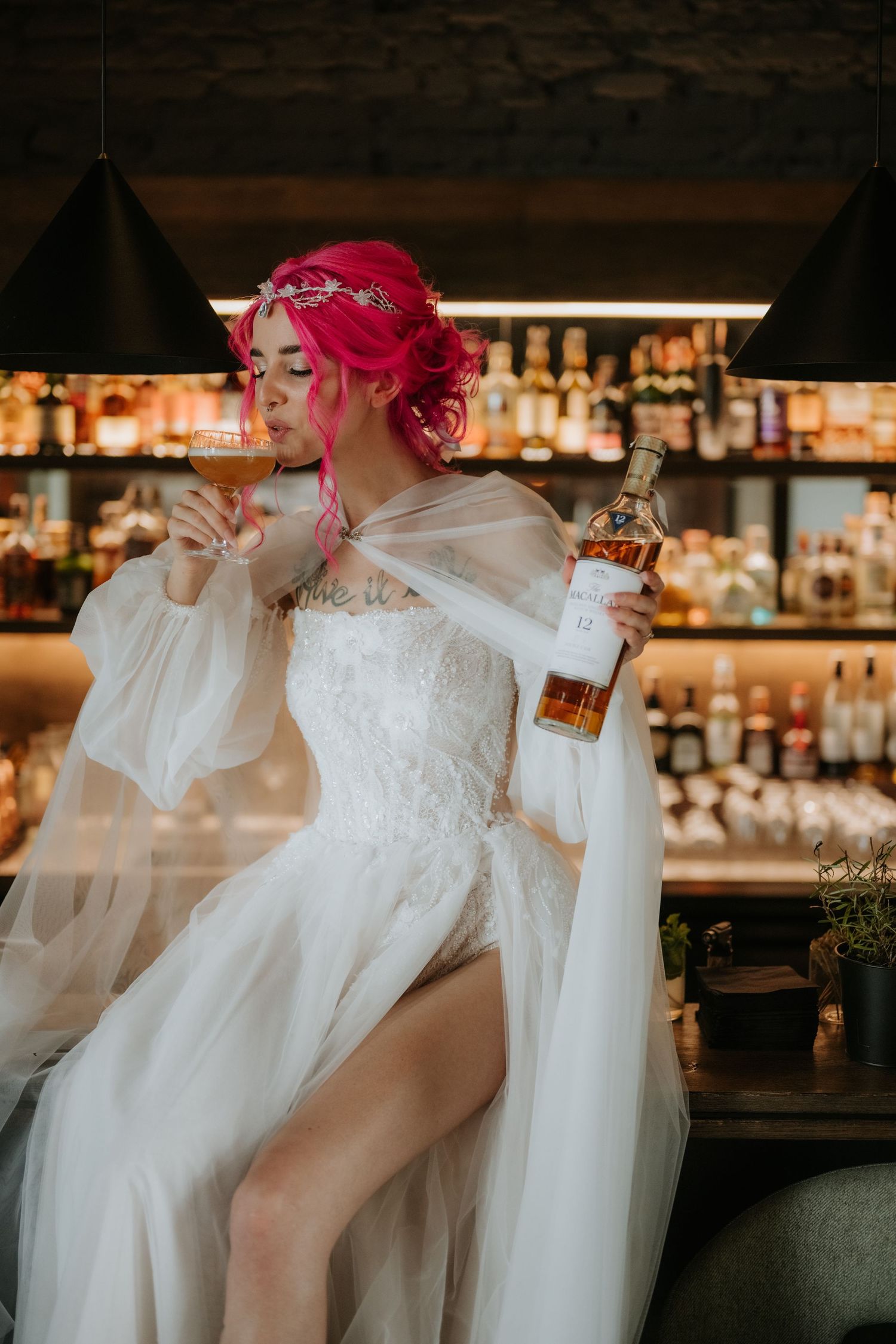 Bride with pink hair in white lace gown holds whiskey bottle and cocktail glass at a bar.