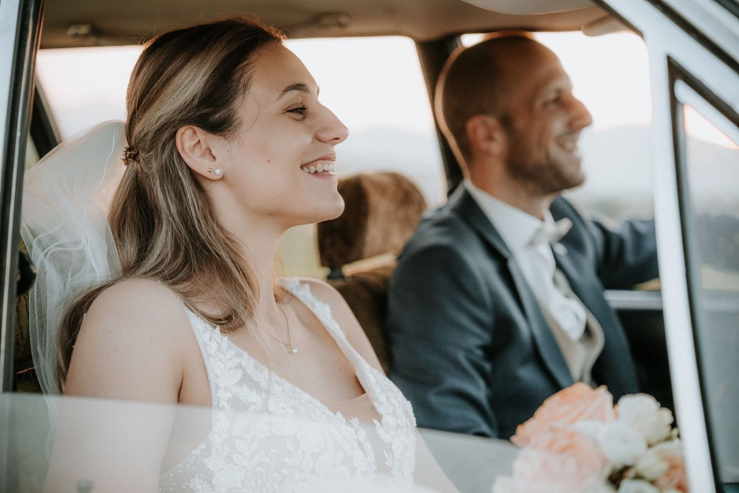Smiling bride in white dress holding bouquet sits beside groom in navy suit inside a car on wedding day.