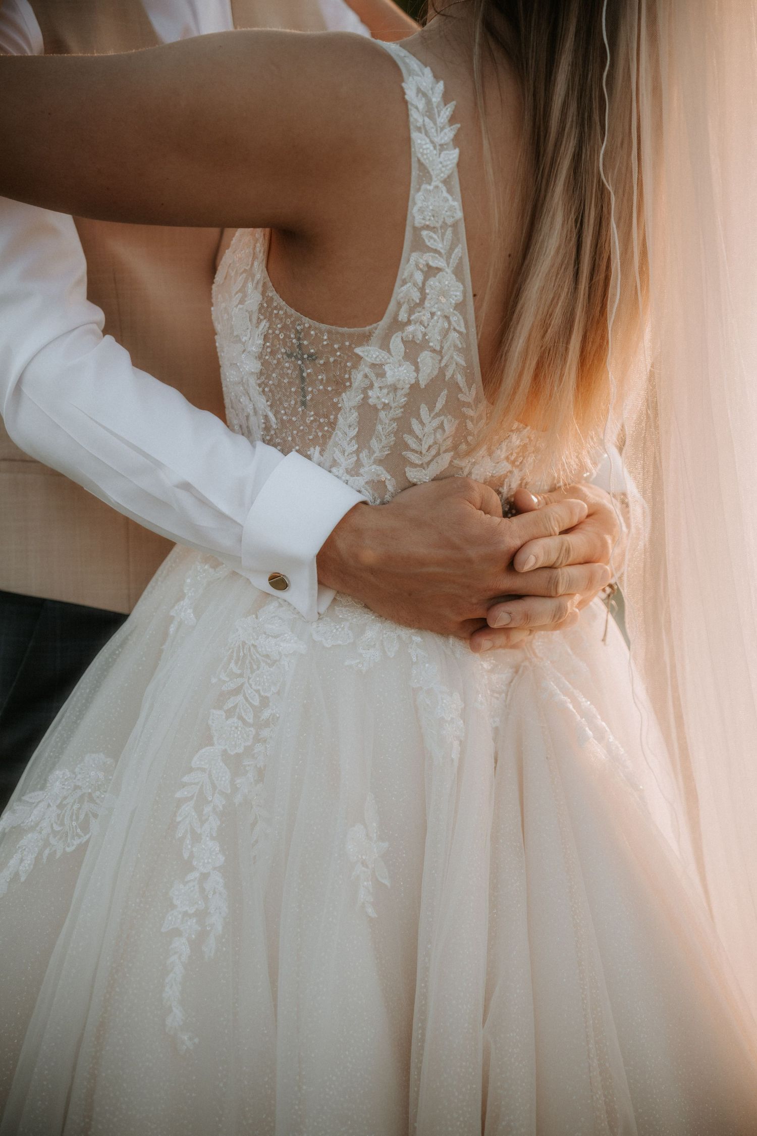 Groom in white shirt embracing bride in lace wedding dress from behind, hands clasped at her waist.