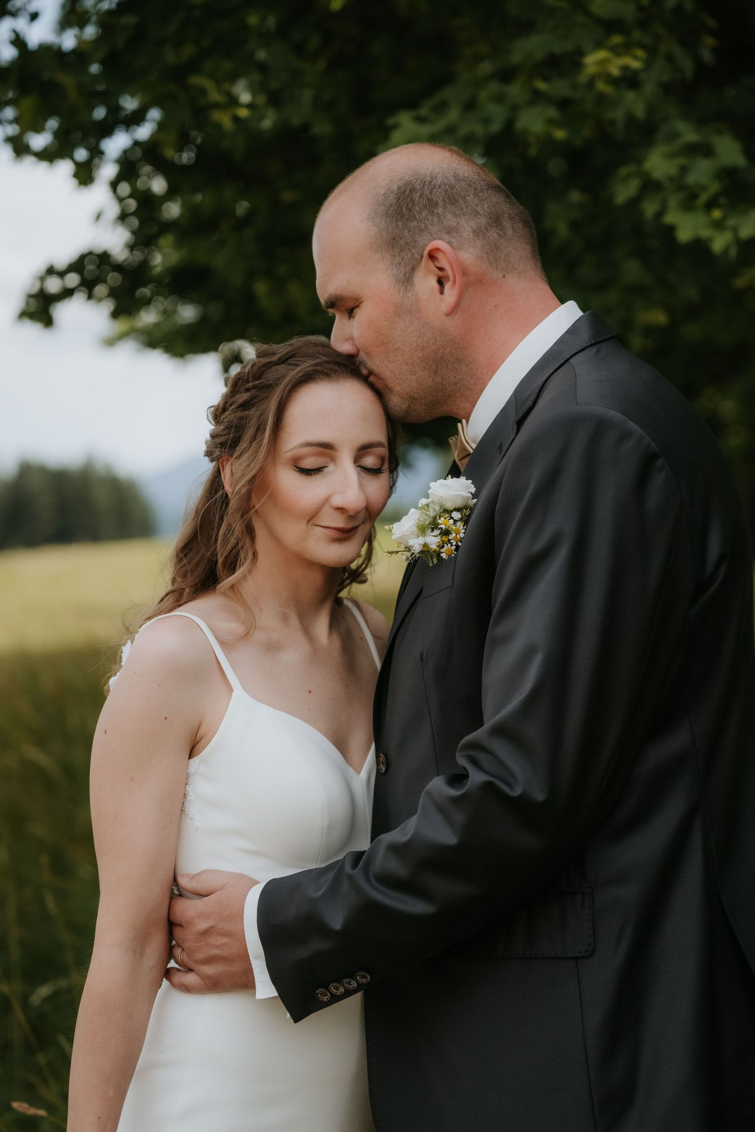 Groom in dark suit kisses bride on temple outdoors, she wears white dress, eyes closed, green trees behind them.