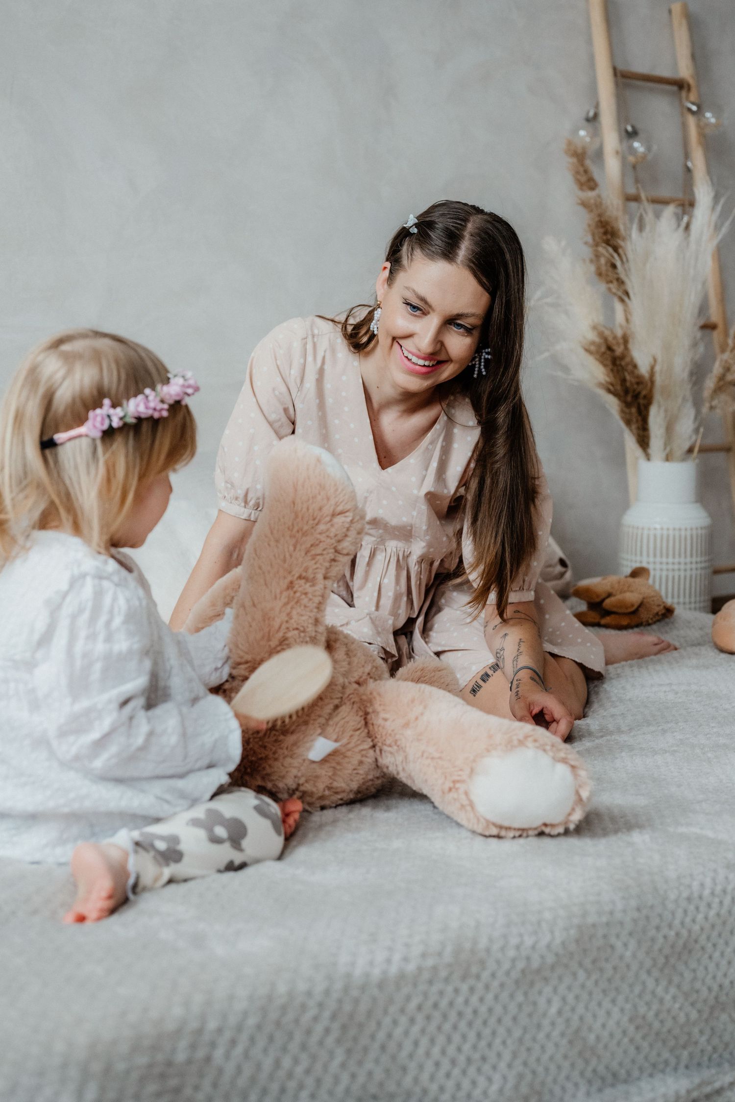 A smiling mother sits on a bed with her toddler daughter sharing a large stuffed teddy bear.