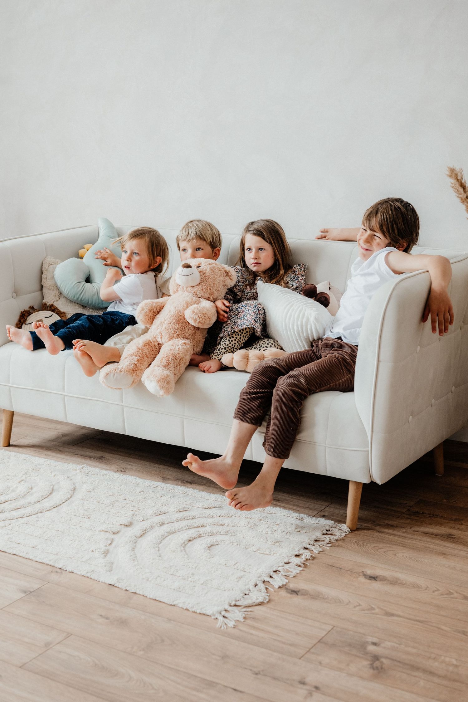 Four young children relaxing together on a beige sofa with stuffed animals and pillows.