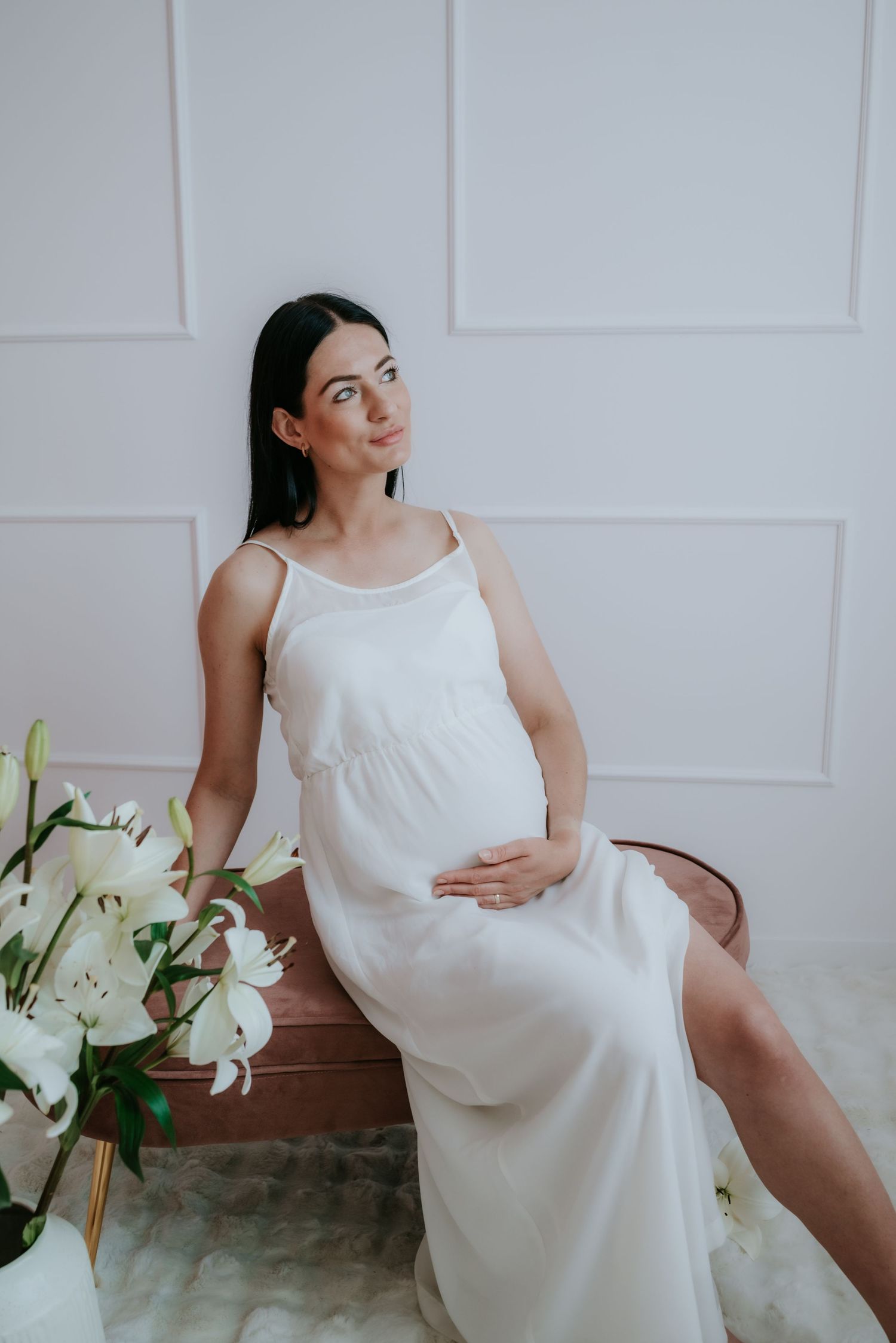 Pregnant woman in white slip dress seated beside white lilies, gazing upward thoughtfully.