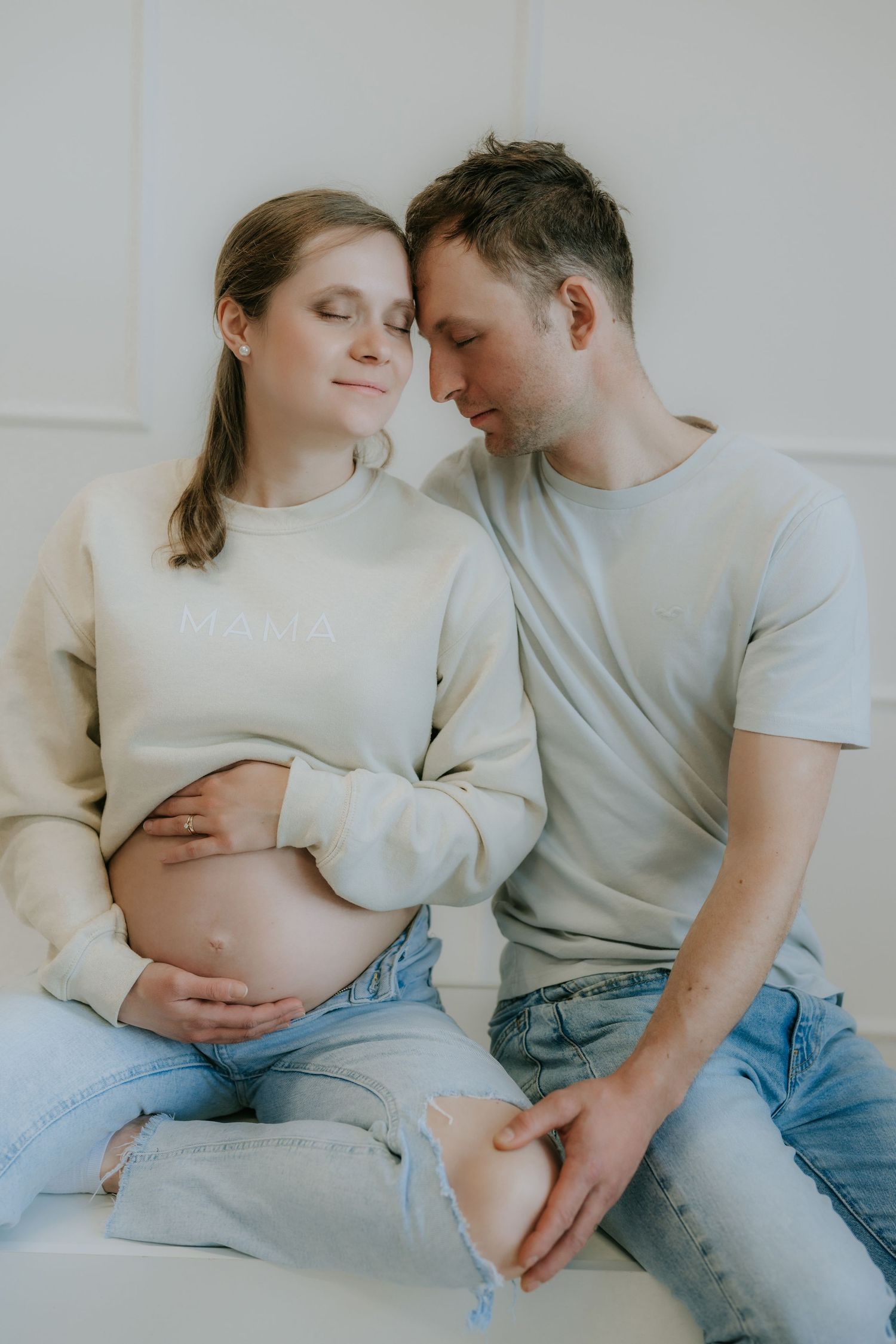 Expecting couple in matching beige outfits sitting close together with eyes closed, maternity shoot.