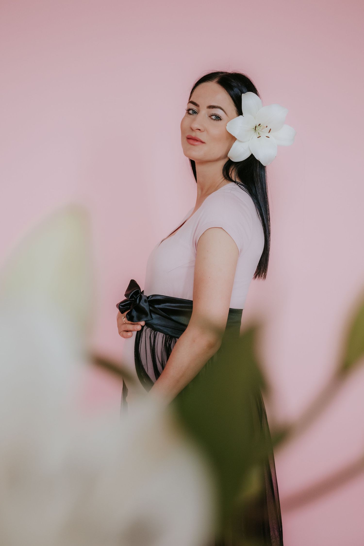 Pregnant woman with white flower in hair wearing pink top and black tulle skirt on pink background.