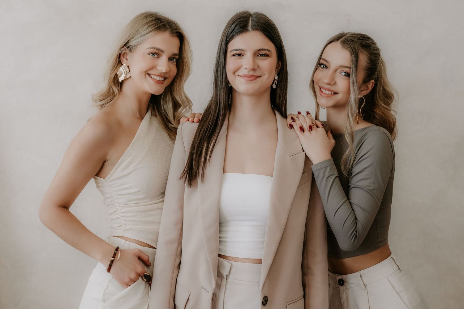 Three young women smiling together in neutral-toned outfits against a light beige background.