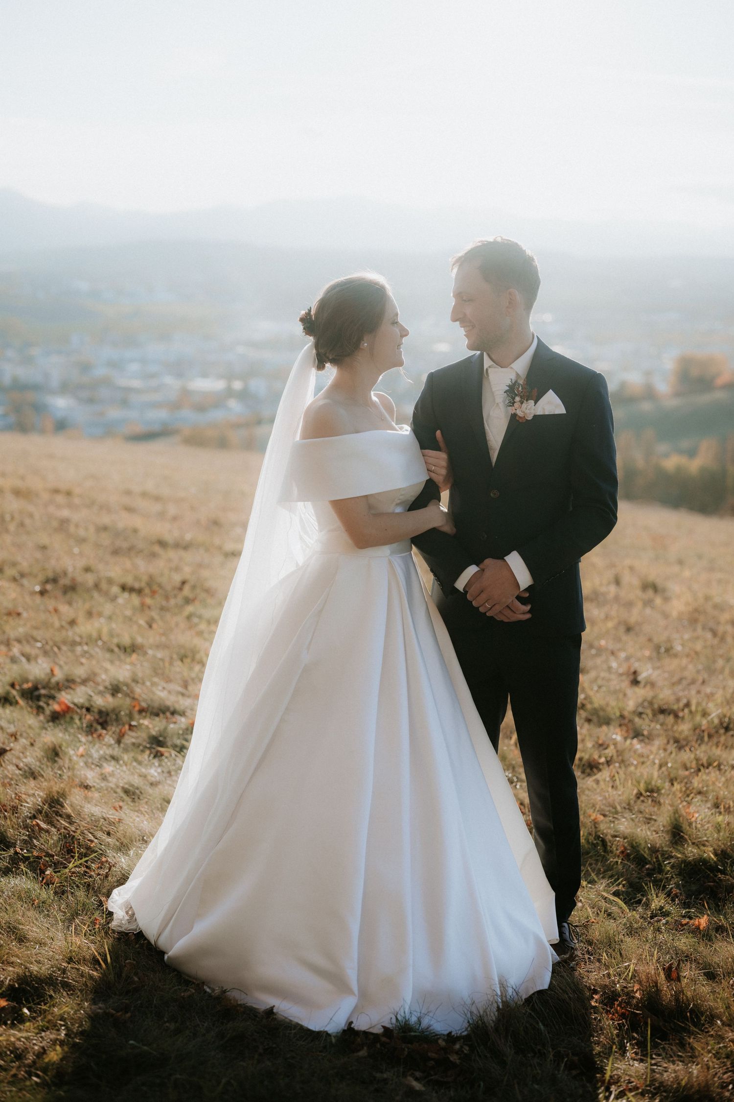 Bride in flowing white off-shoulder gown and veil standing with groom in black suit on a golden hillside.
