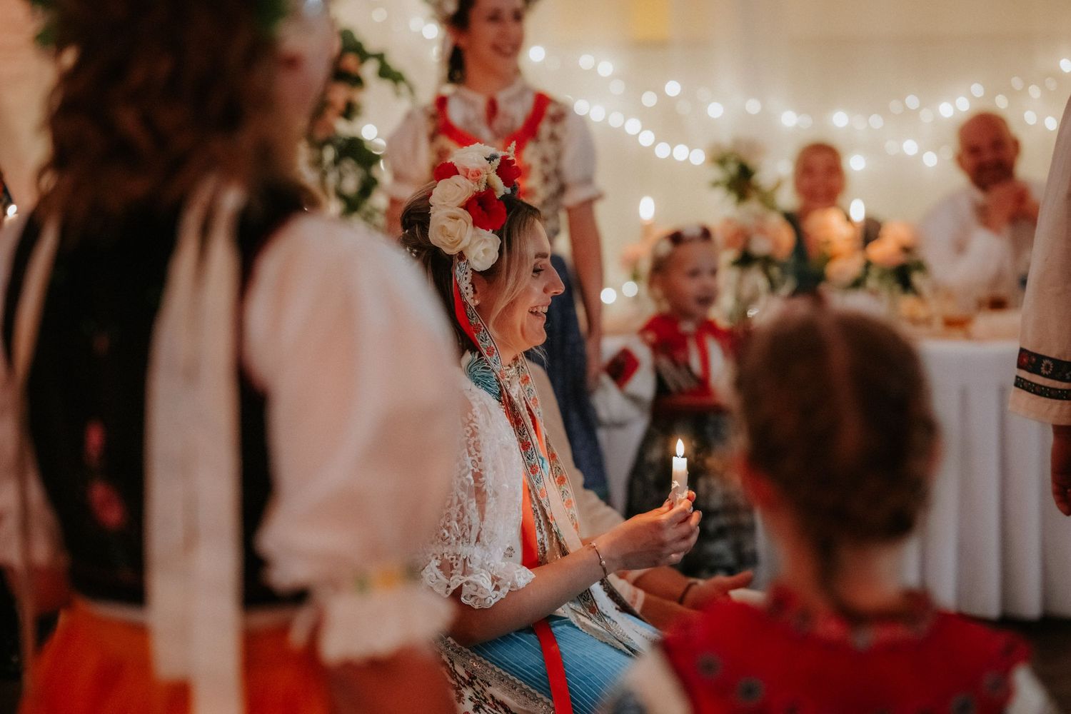 Woman in traditional folk costume holding candle at festive cultural celebration with fairy lights.