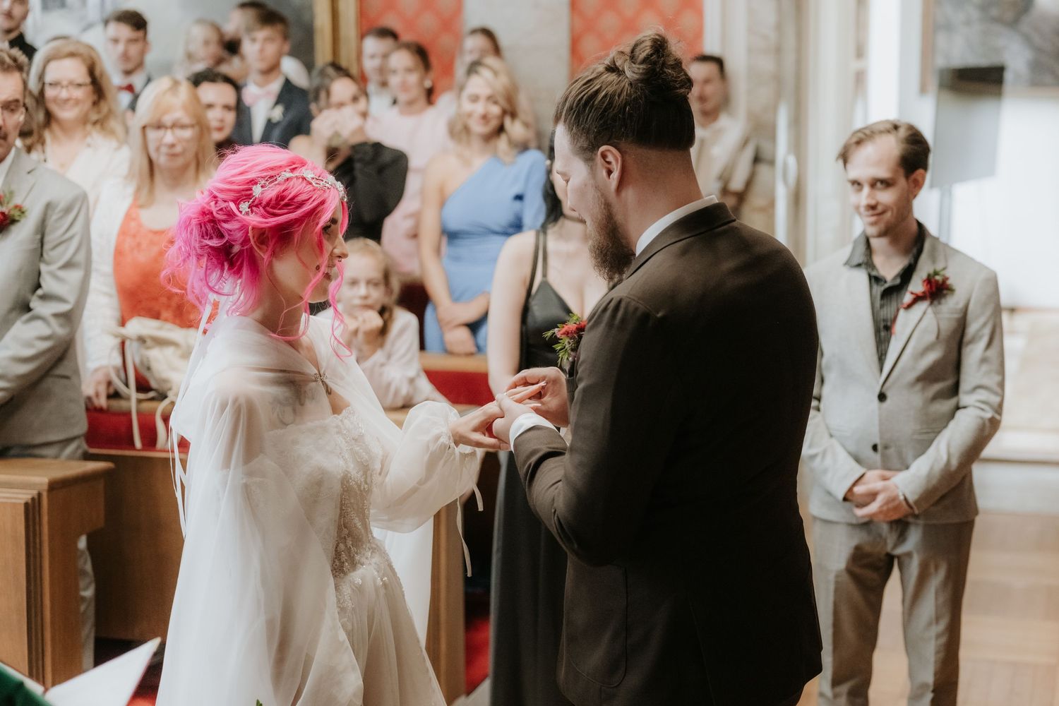 Bride with pink hair exchanges rings with groom during wedding ceremony, guests watching in elegant venue.
