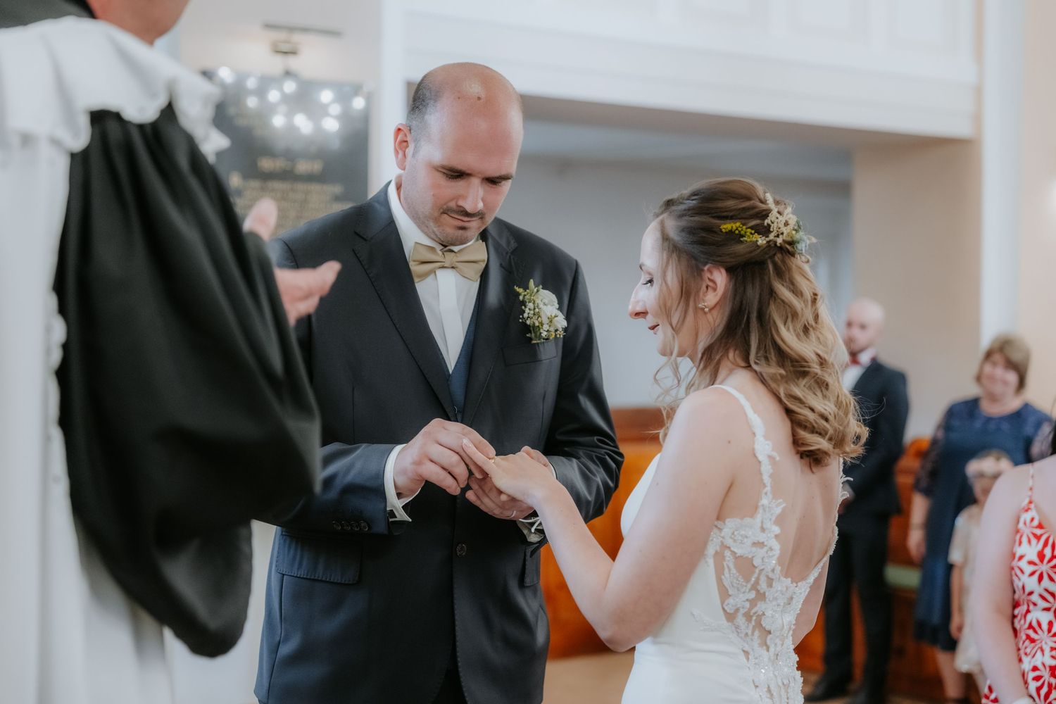 Groom placing wedding ring on bride's finger during ceremony, wearing navy suit with gold bow tie.