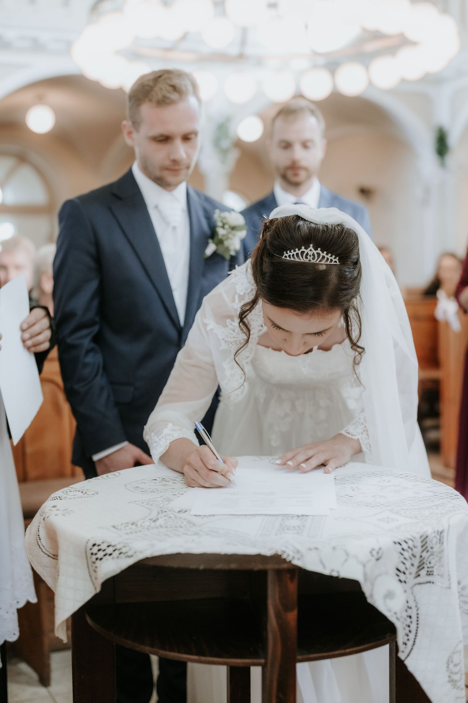 Bride in white gown and tiara signing marriage certificate at church altar with groom in navy suit standing behind.