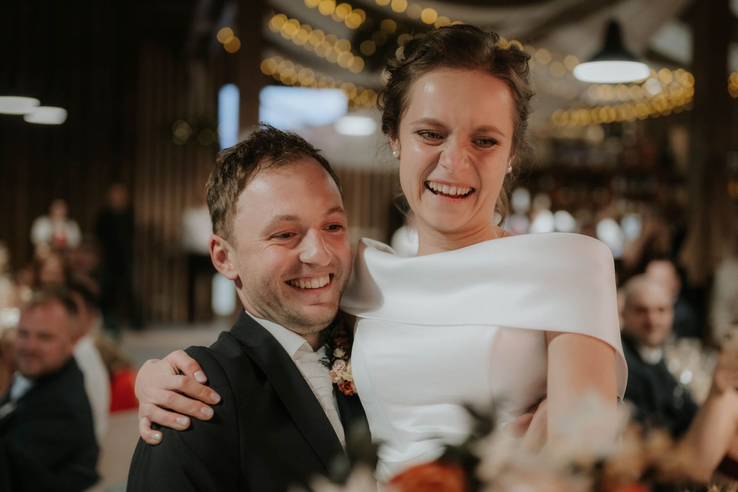 Happy couple embracing and laughing at their wedding reception in a warmly lit barn venue.