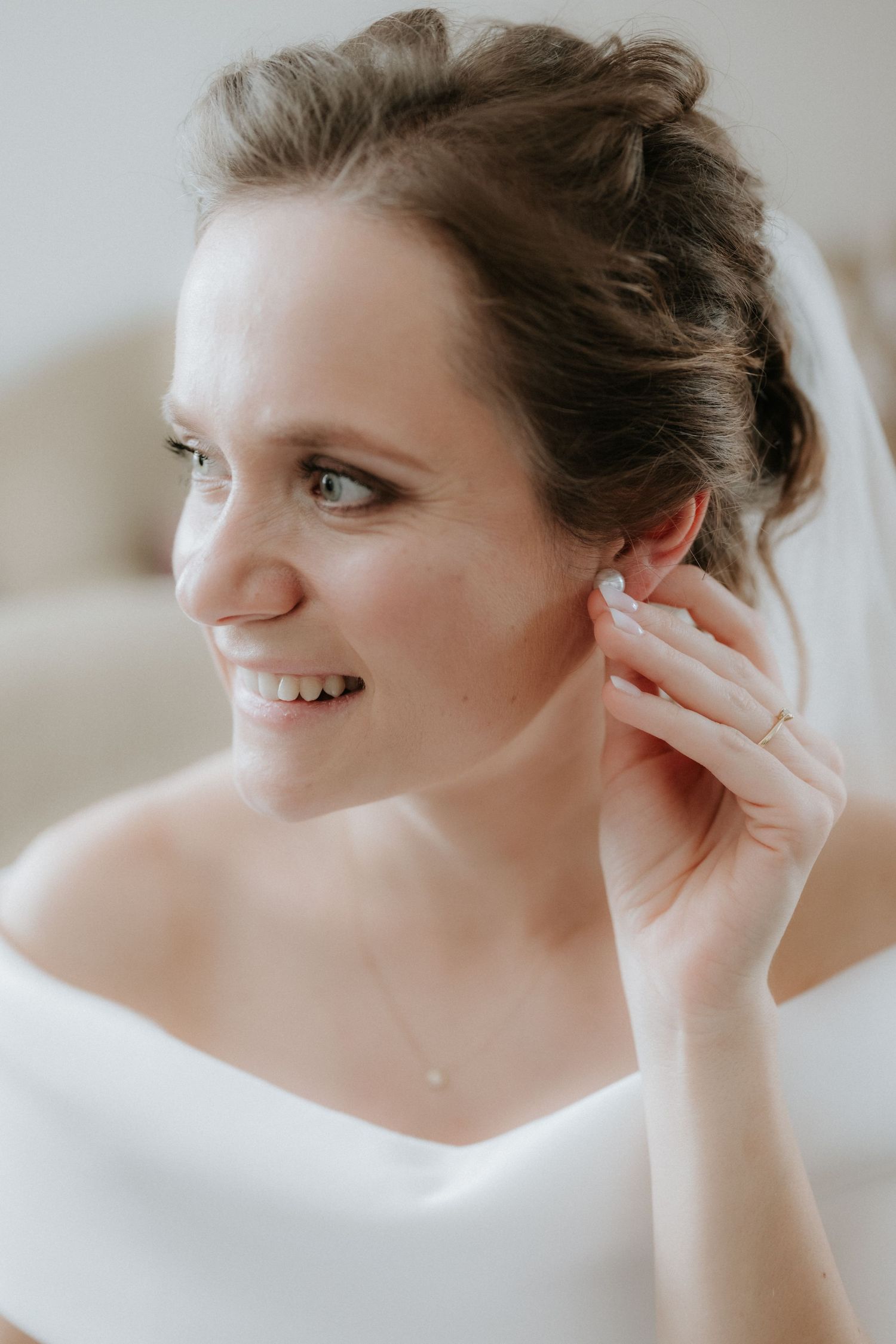 Smiling bride in white off-shoulder gown adjusting her earring, with an elegant updo and veil.