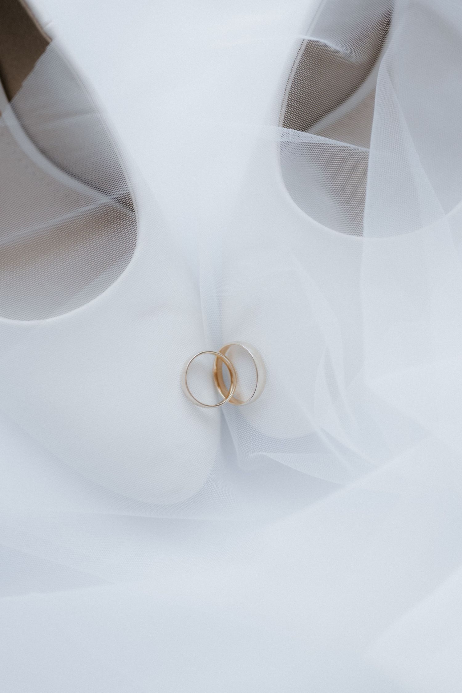 Two gold wedding rings rest on a white bridal veil beside elegant white high heel shoes.