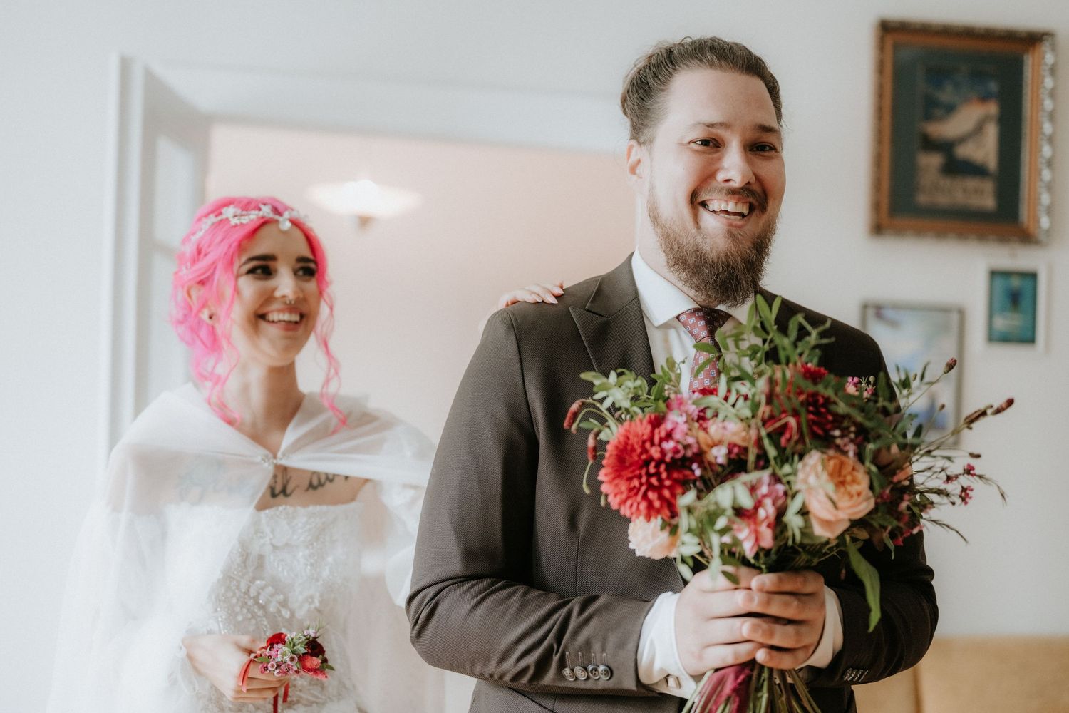 Smiling groom holds colorful bridal bouquet with pink-haired bride in white dress and cape standing behind him.