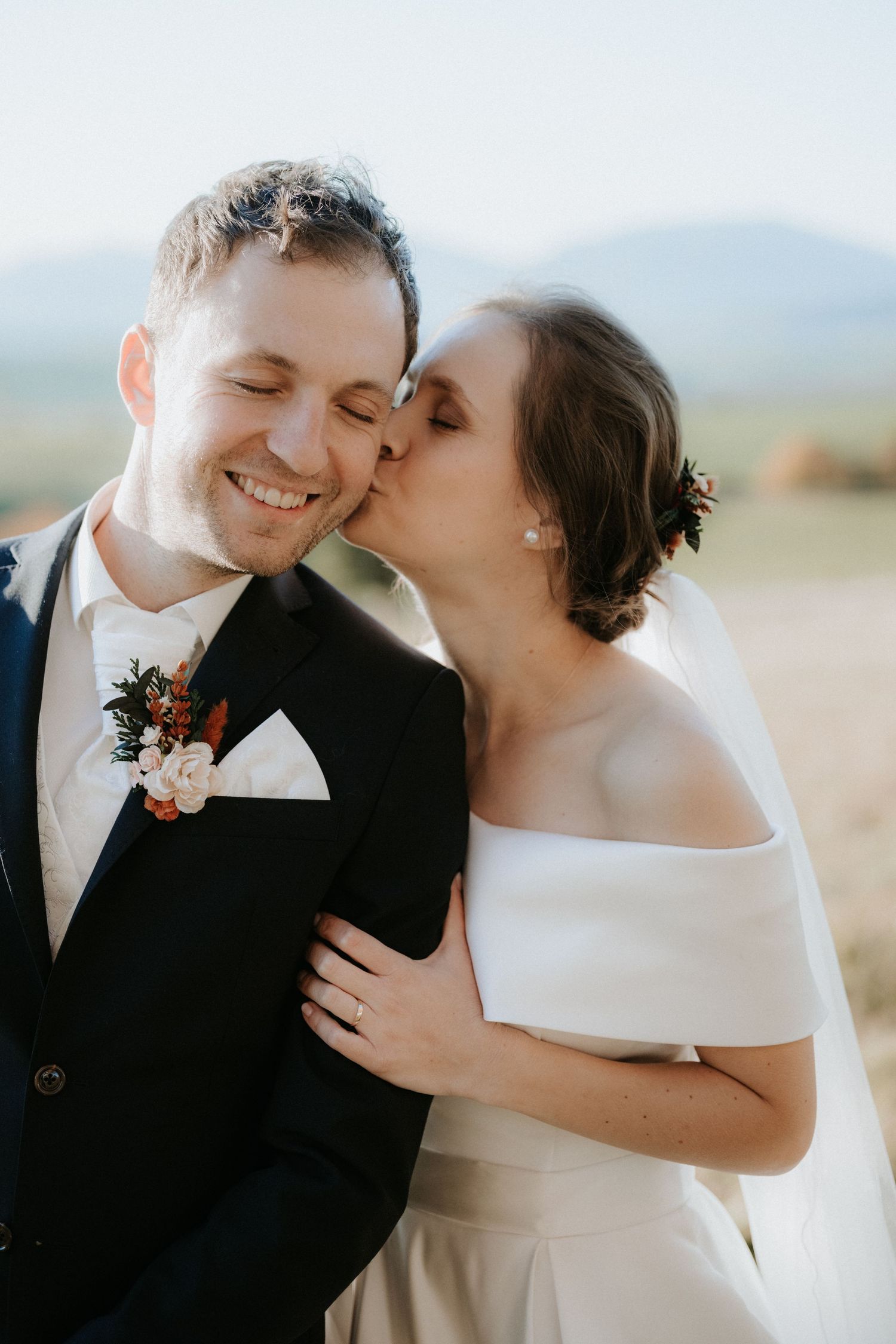 A bride in an off-shoulder white gown kisses her smiling groom in a navy suit with floral boutonniere outdoors.