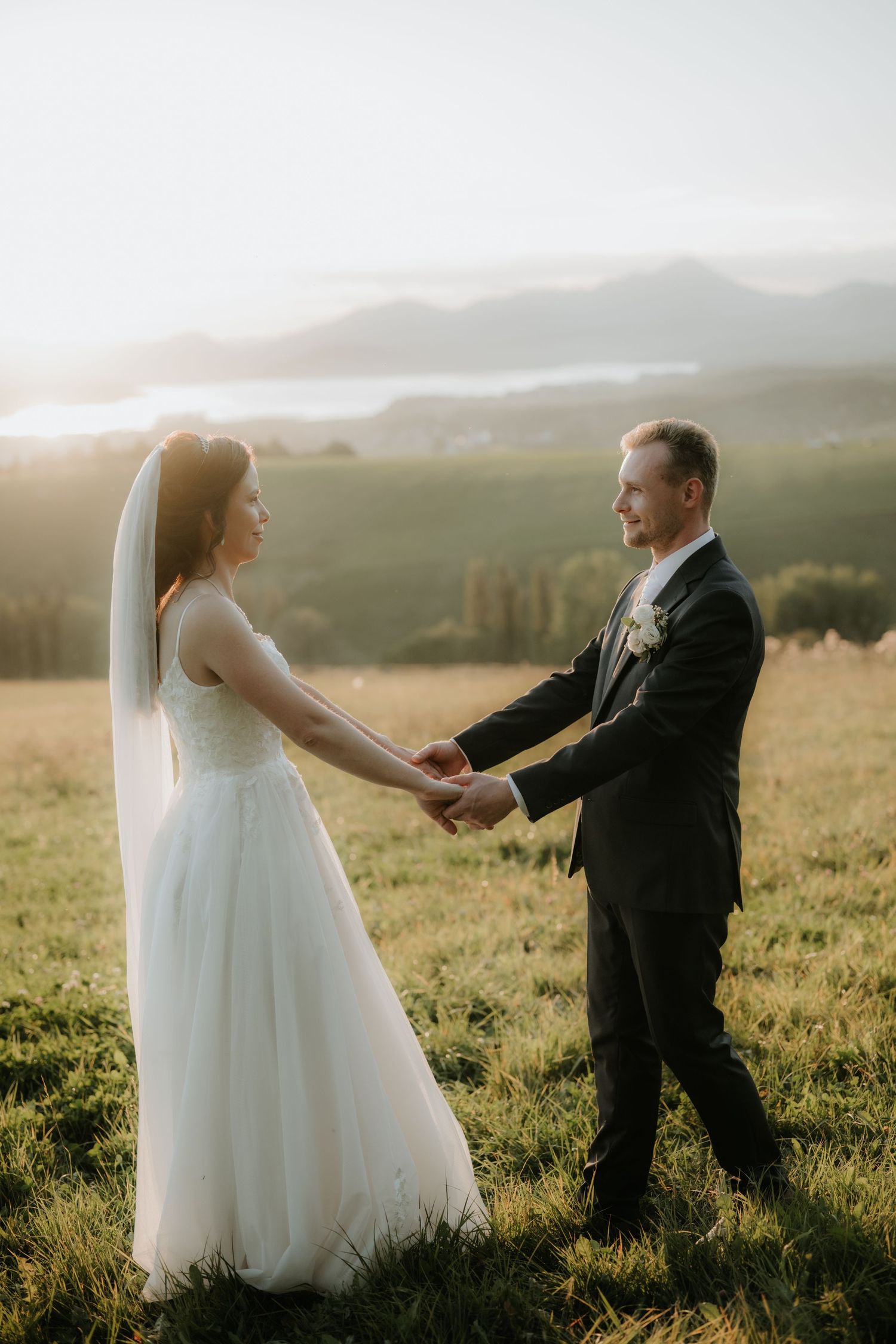 Bride and groom holding hands in a sunlit meadow with misty mountains and a lake in the background.