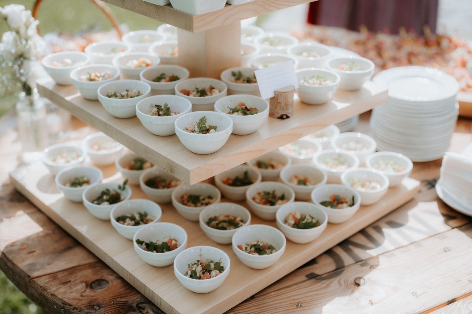 Tiered wooden display stand holding multiple white bowls filled with appetizers at an outdoor catering event.