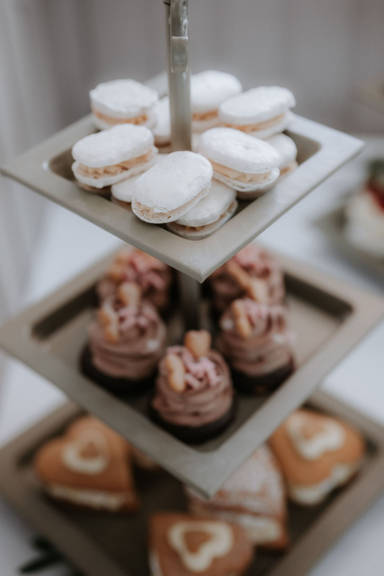 Three-tier dessert stand displaying white macarons, frosted cupcakes, and heart-decorated pastries at an elegant event.