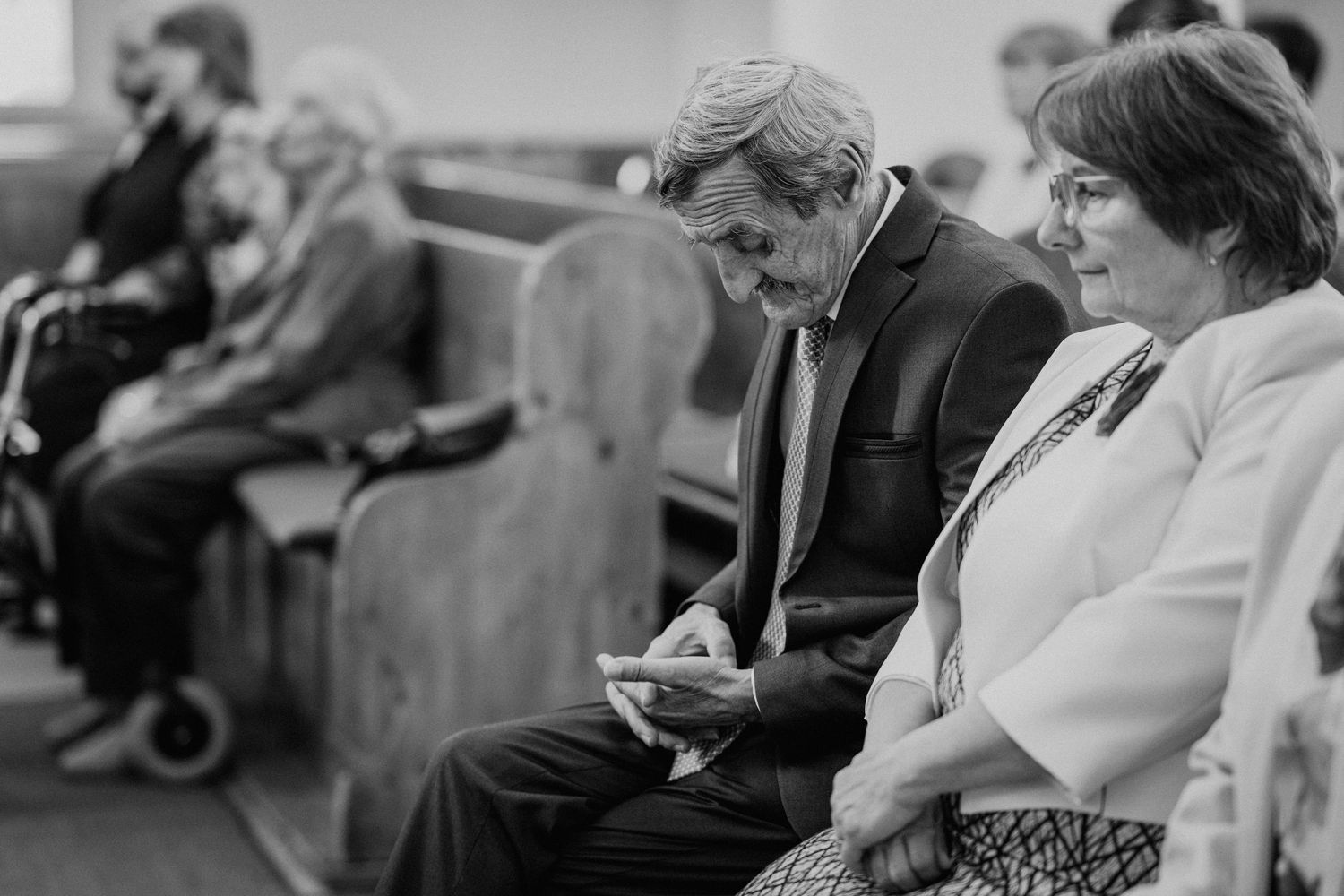 Black and white photo of guests seated in church pews during a wedding ceremony, man in suit reviewing notes.
