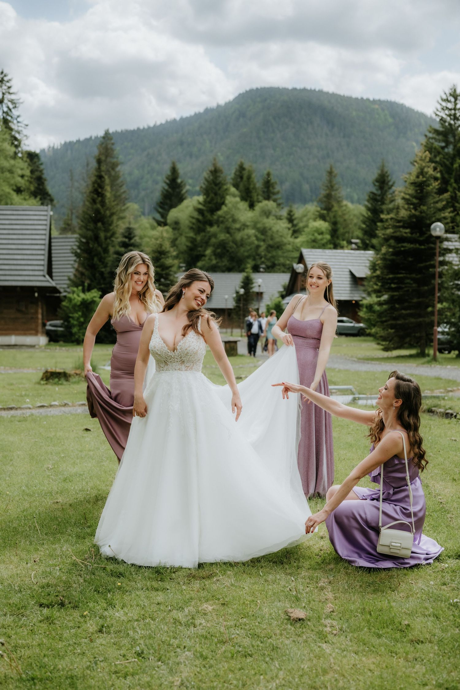 Bride in white gown with three bridesmaids in purple dresses outdoors, mountains and pine trees in background.