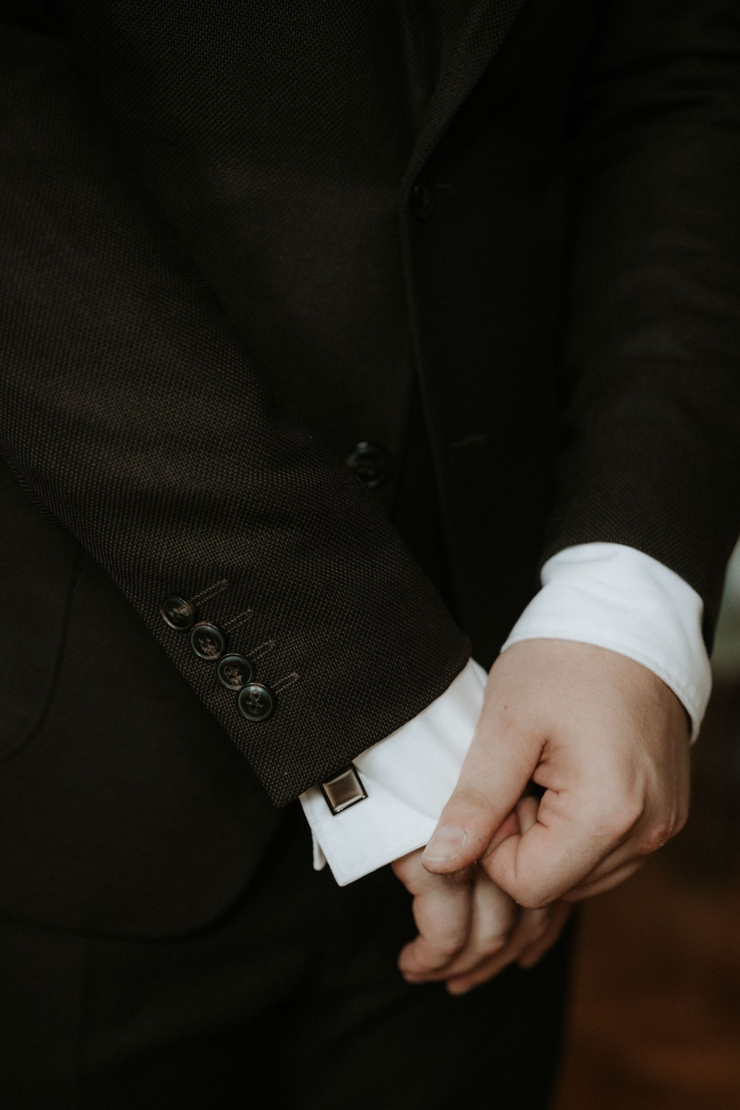 Close-up of a man in a dark suit with white dress shirt cuff and gold cufflink, hands clasped together.
