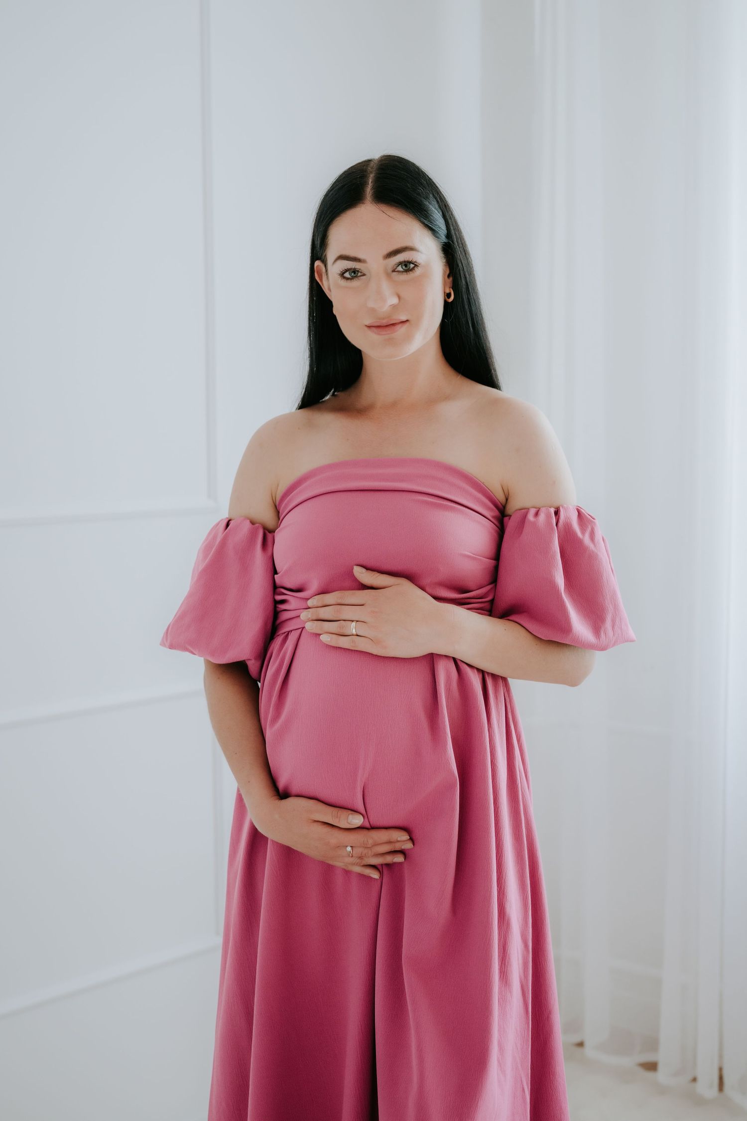 Pregnant woman in pink off-shoulder dress cradling baby bump in bright white room.