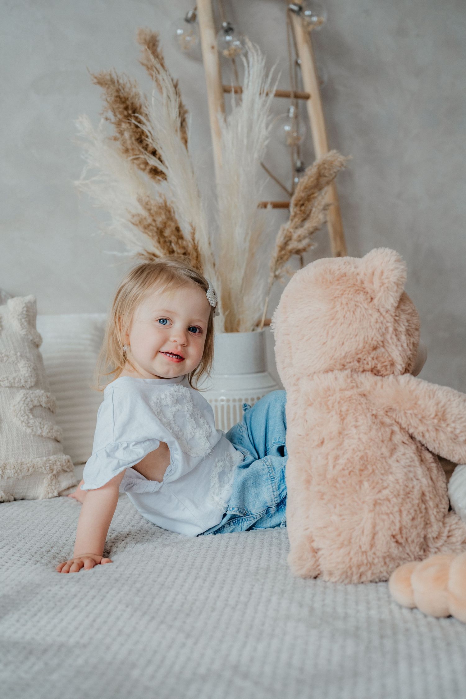 Cute blonde toddler girl in white top and jeans sitting on bed with large teddy bear and pampas grass decor.