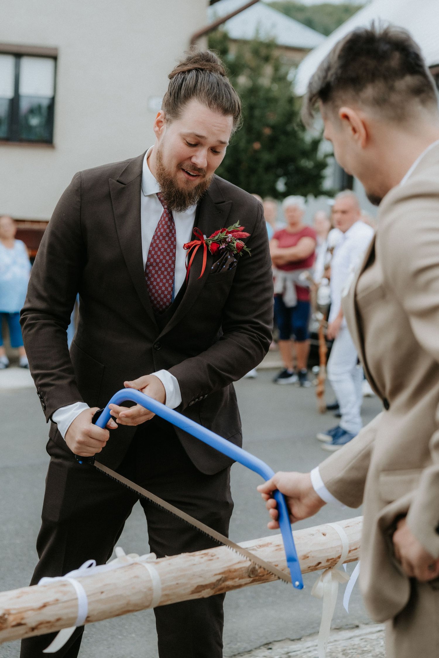 Groom in dark suit with boutonniere sawing a log with another man in a traditional wedding ceremony outdoors.