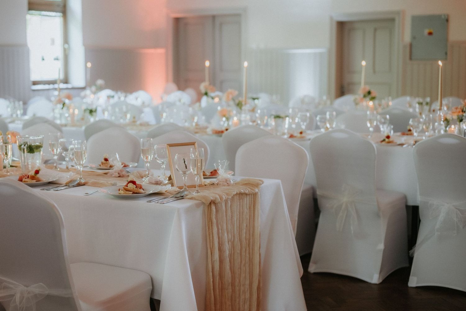 Elegant wedding reception hall with white draped chairs, candlelit tables, and gold accents in a classic venue.