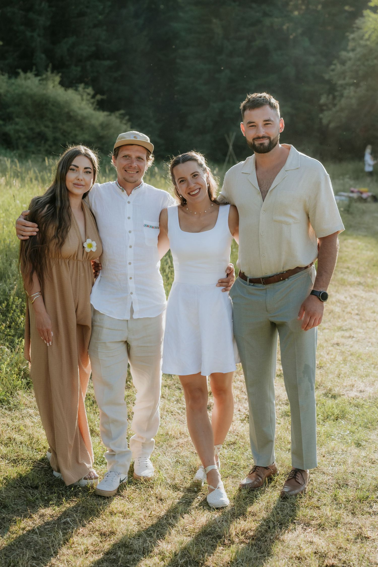 Four friends smiling outdoors in summer attire, posing together on grass with green trees in background.