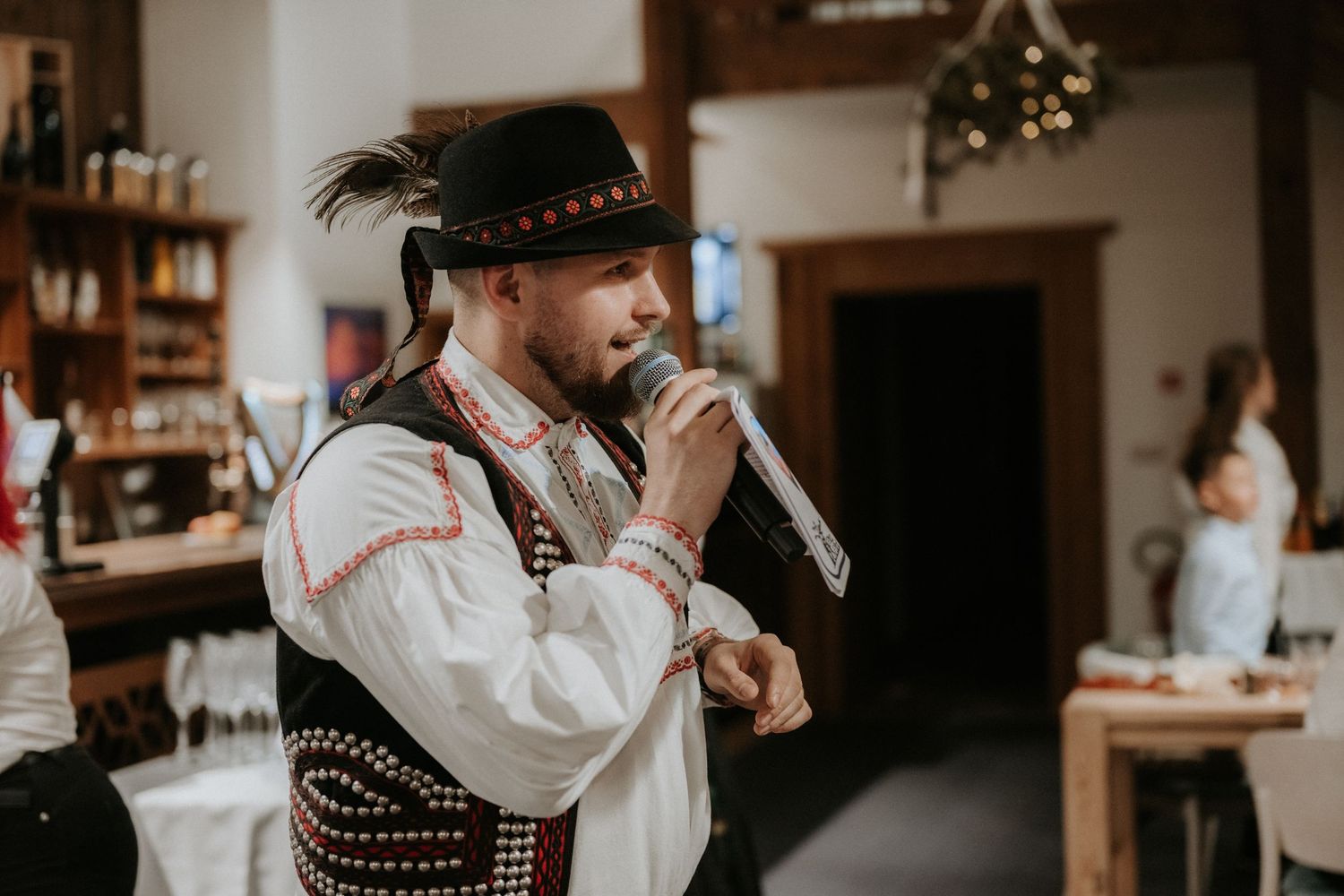 Man in traditional folk costume and feathered hat singing into microphone at indoor cultural event.