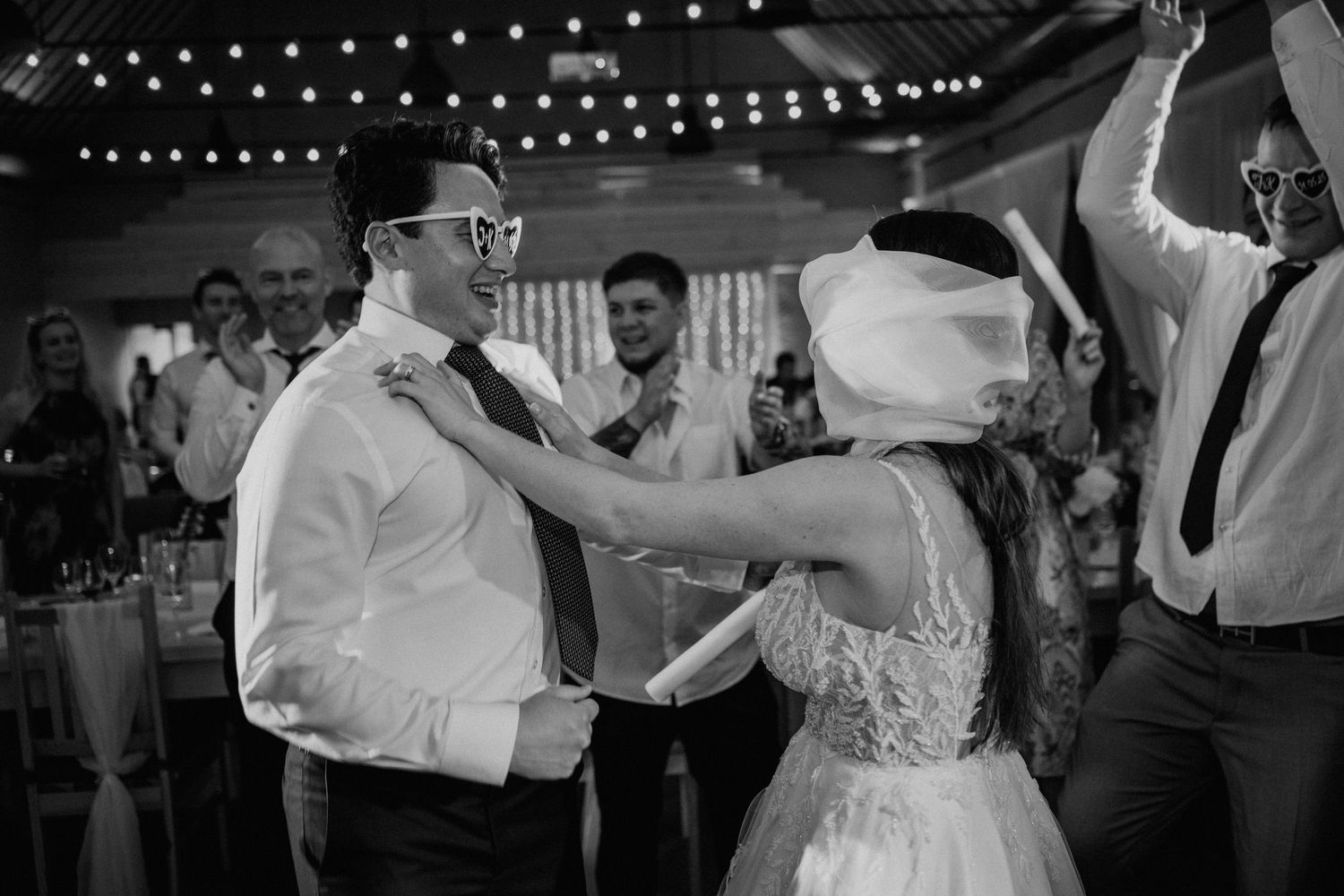Black and white photo of a bride and groom dancing joyfully at their wedding reception with guests celebrating in background.