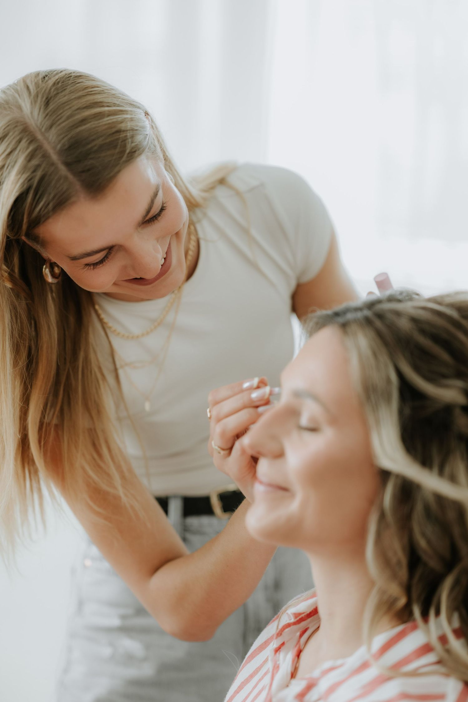 A smiling makeup artist applies makeup to a relaxed woman with closed eyes in a bright, airy studio setting.
