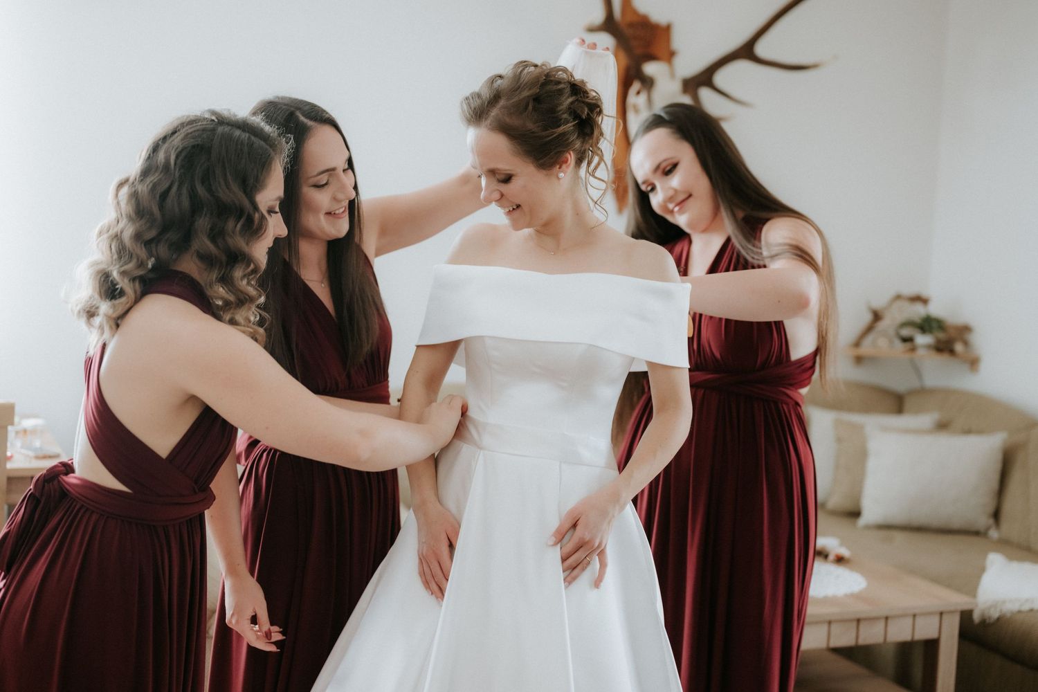Bride in white off-shoulder gown assisted by three bridesmaids in burgundy dresses during wedding preparation.