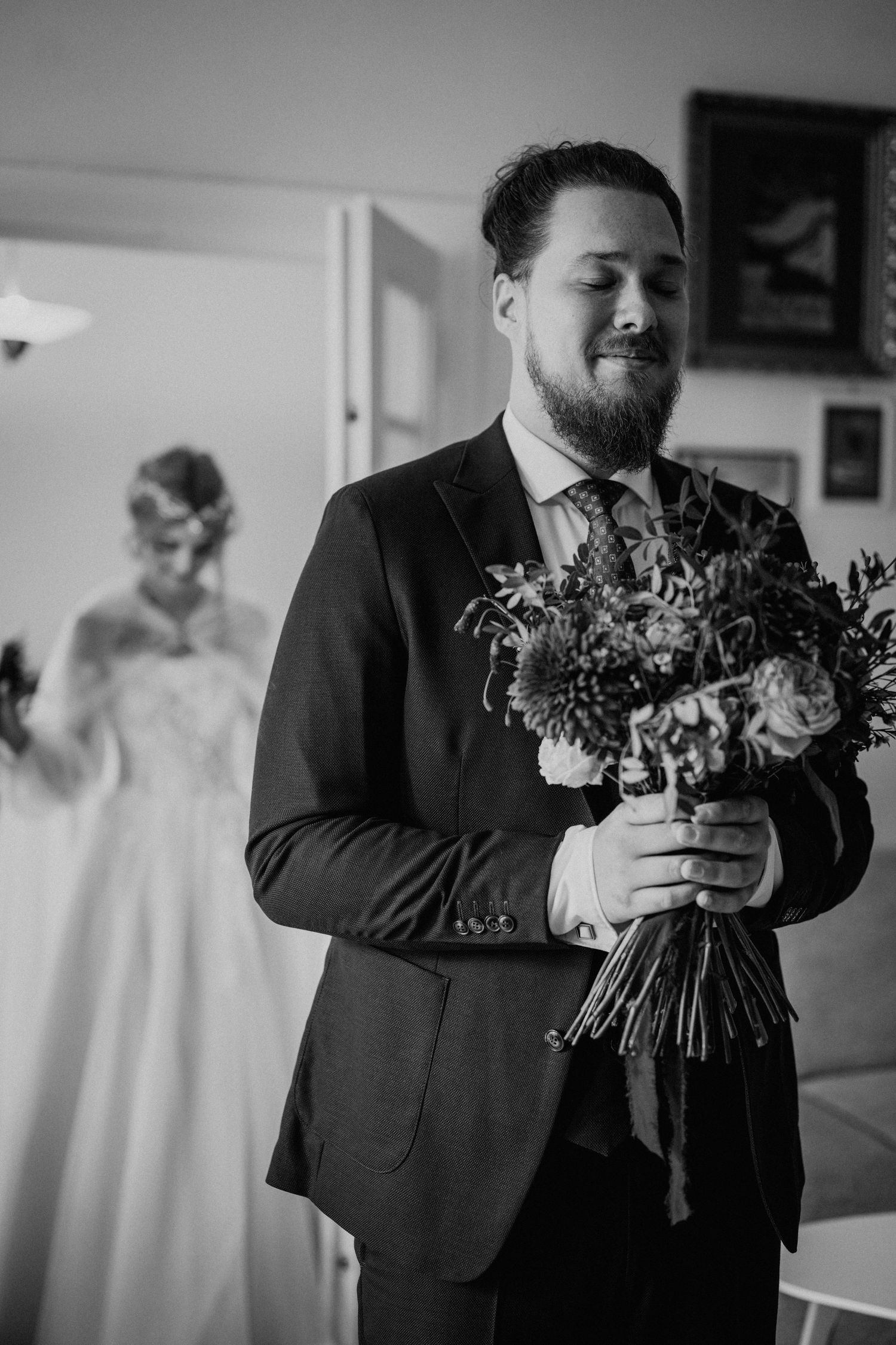Black and white wedding photo of a bearded groom holding a bouquet with the bride standing in the background.